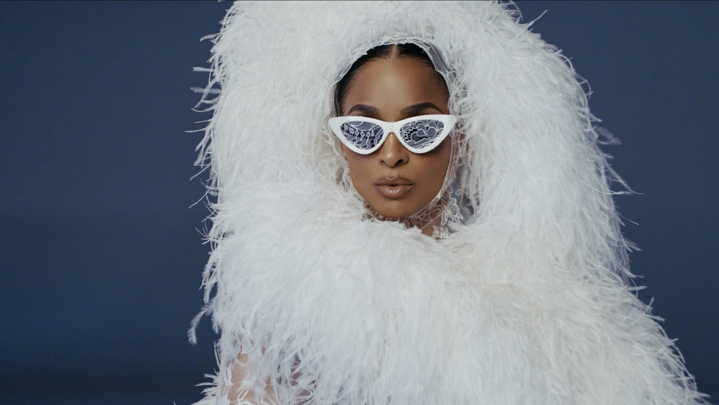 A woman wearing white fuzzy coat, large white feathered headdress, and white sunglasses with decorative lenses, posing against a dark background.