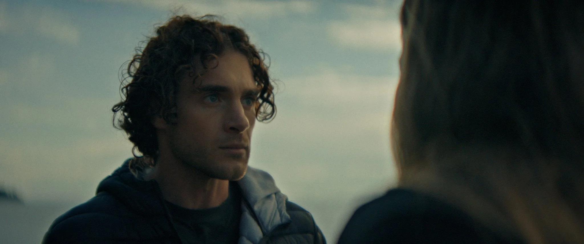 A man with curly hair and intense blue eyes looking at a woman whose face is partly obscured, against an overcast sky background.
