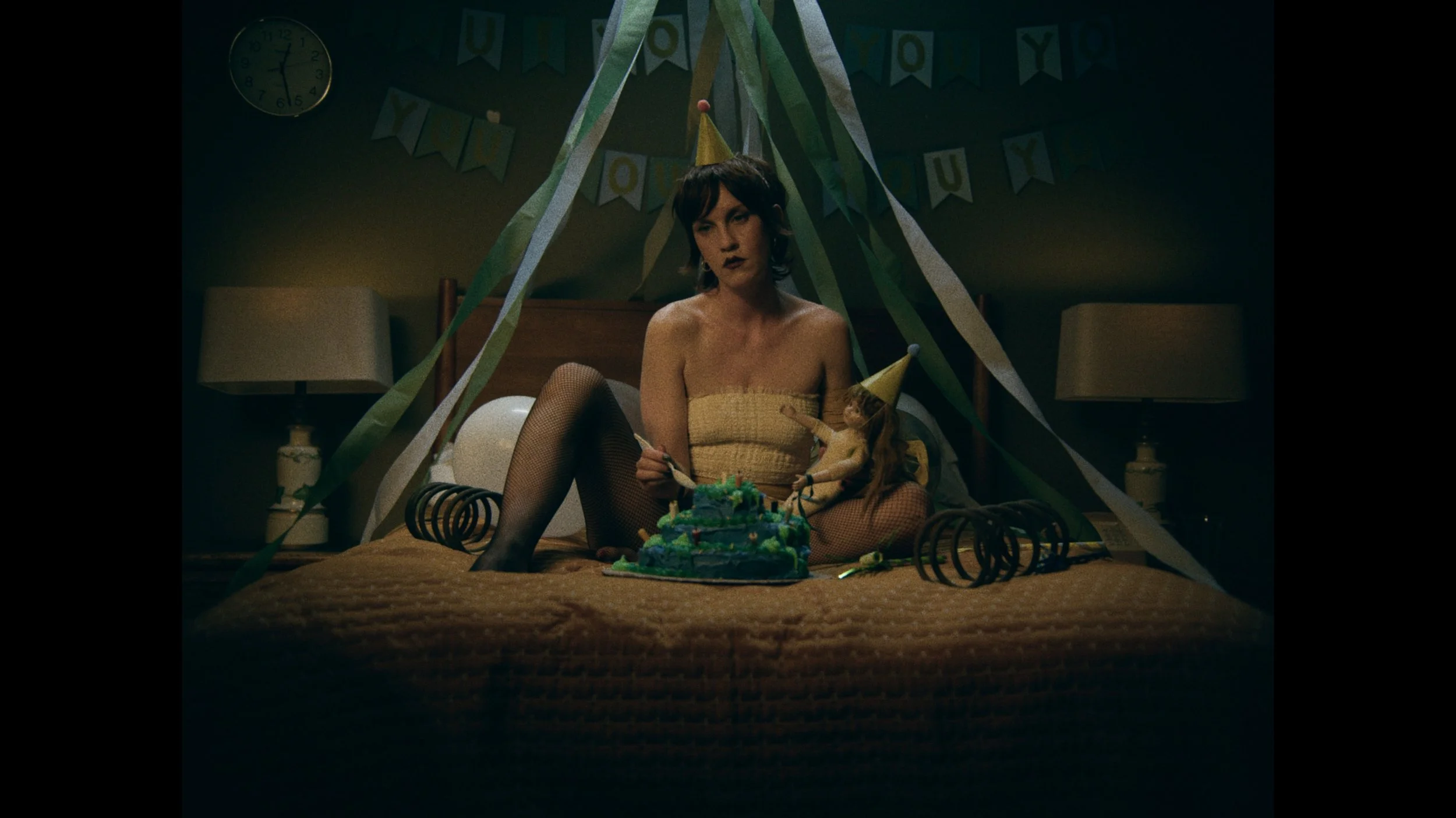 A woman in yellow attire sitting on a bed decorated for a birthday party. She is wearing fishnet stockings and a birthday hat, holding a cake or object, with party streamers and a doll as decorations around her.