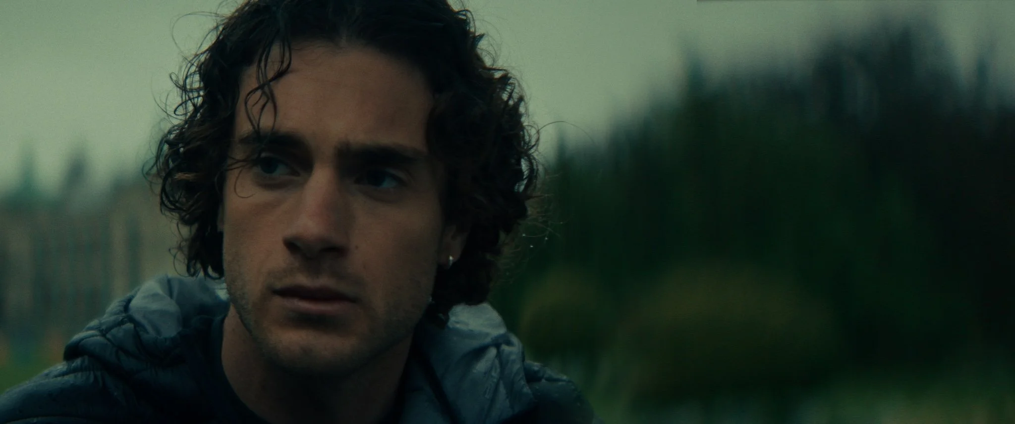 Close-up of a young man with curly dark hair and an earring, looking off to the side with a serious expression, outdoors with a blurred natural background