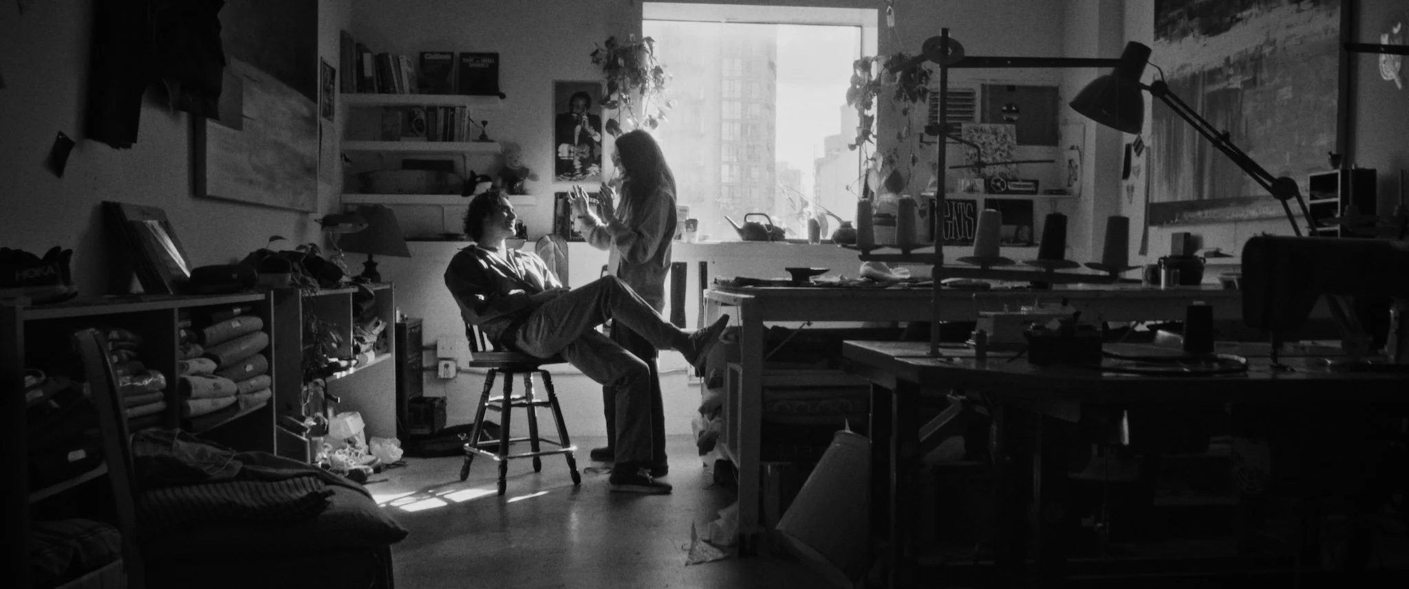 Silhouette of two people in a cluttered, sunlit room, one sitting on a chair with legs crossed and the other standing, engaged in conversation near a large window.