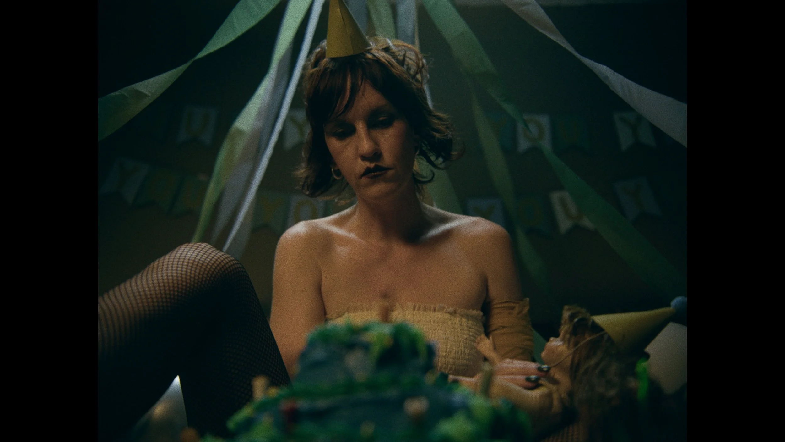 A woman with short, wavy hair, dark makeup, and fishnet stockings celebrating a birthday, wearing a gold party hat, sitting at a table with a decorated cake and a cat wearing a matching party hat, in a dimly lit room with birthday banners.