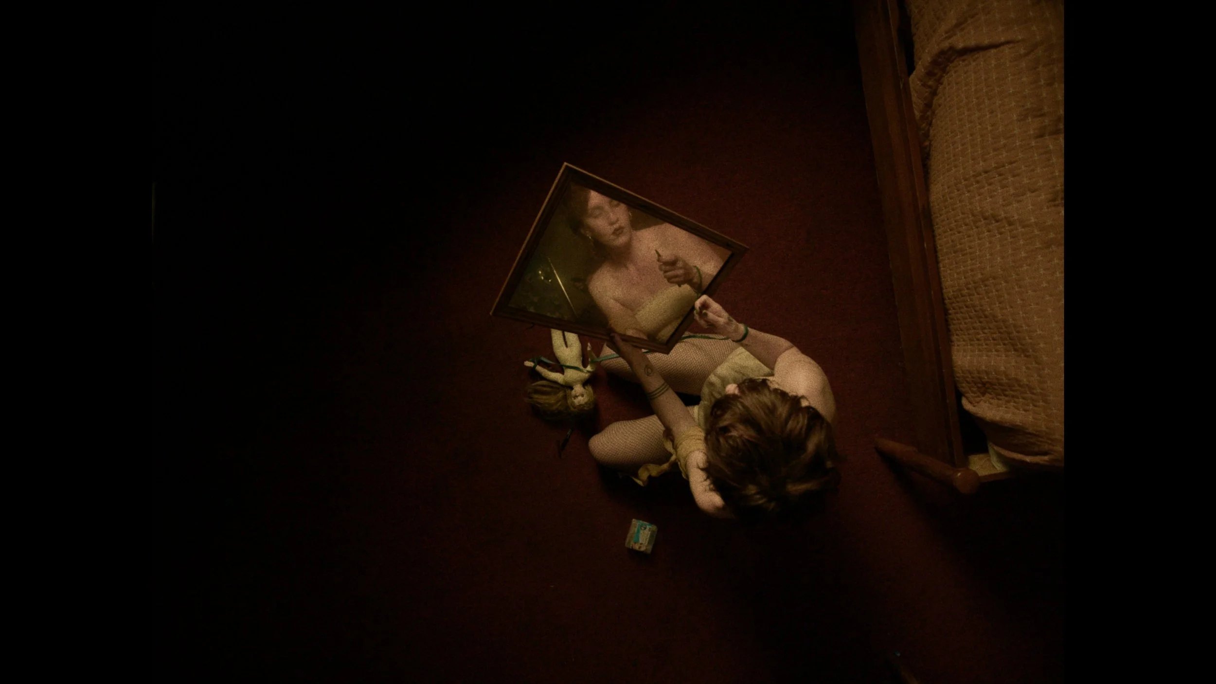 A person lying on the floor, taking a photo of a framed picture that reflects their face, with a bed and a small object nearby.