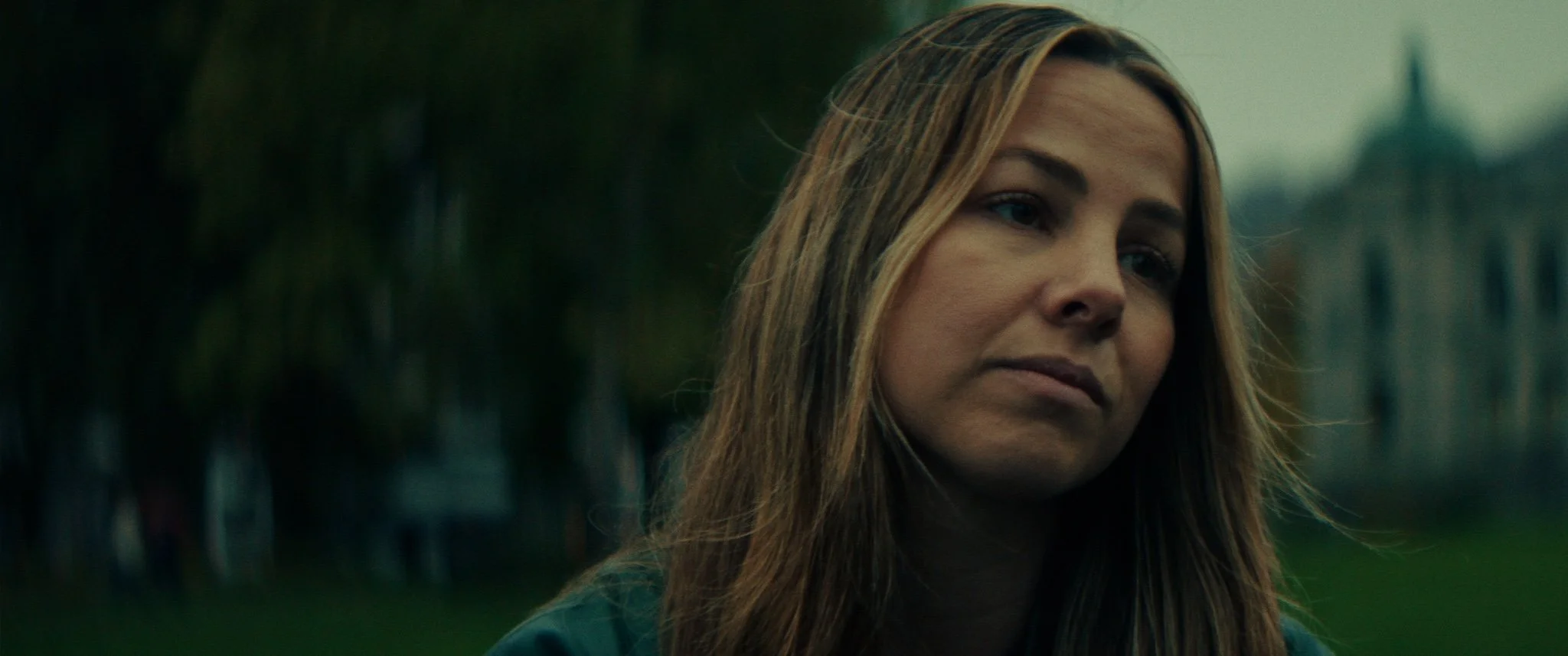 A woman with long brown hair looking to the side with a somber expression, outdoors with blurred trees and buildings in the background.