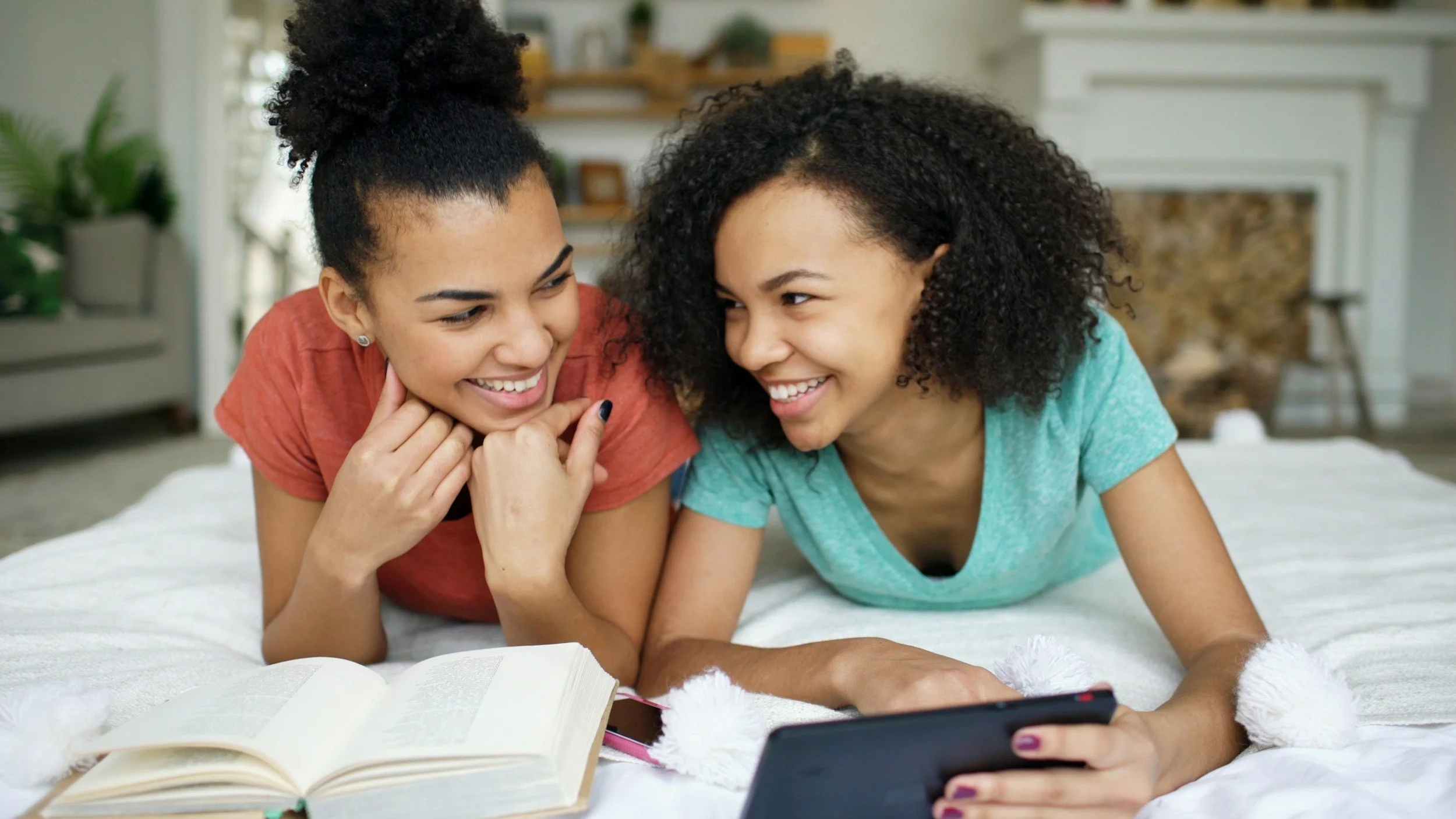 Two young women smiling and looking at a smartphone together while lying on a bed, with an open book nearby.
