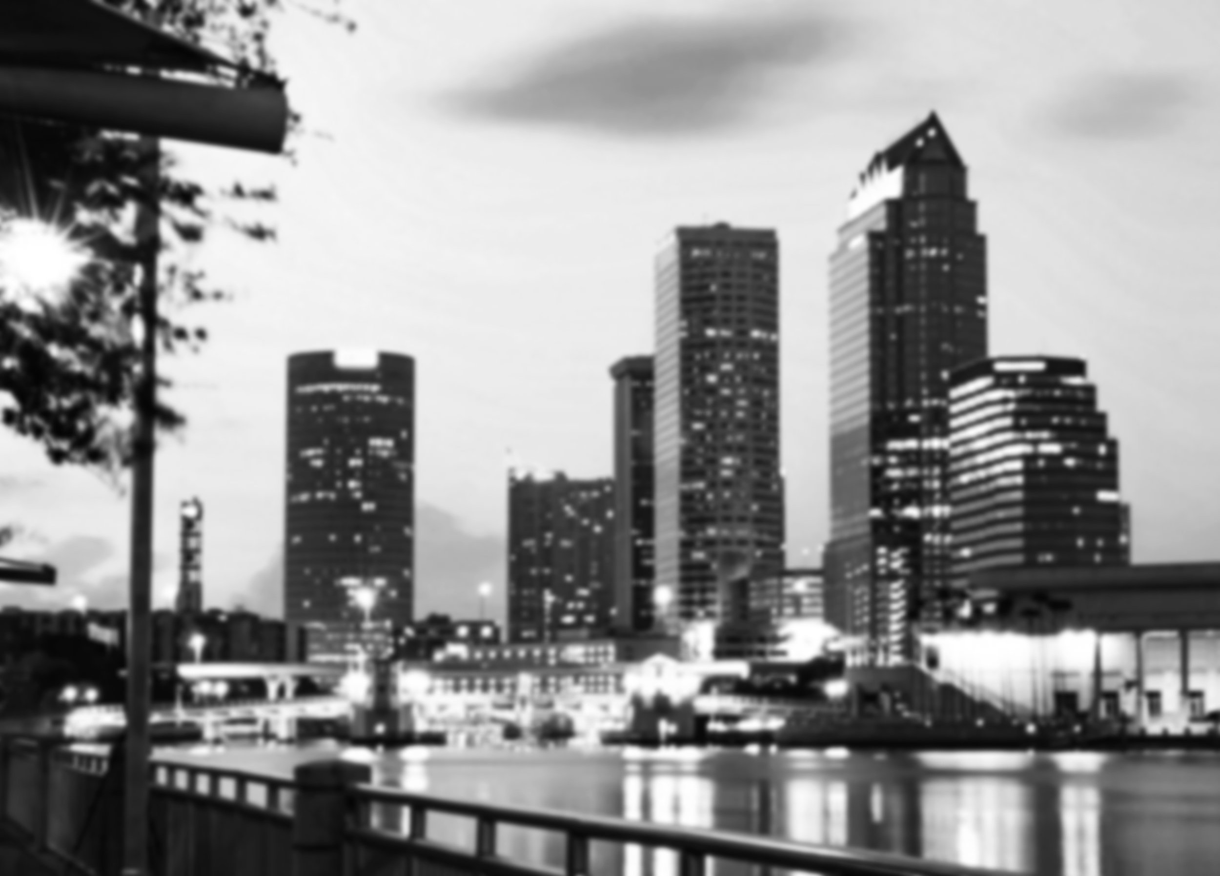 Black and white photo of a city skyline with tall modern buildings, reflecting in water, with a railing and trees in the foreground.