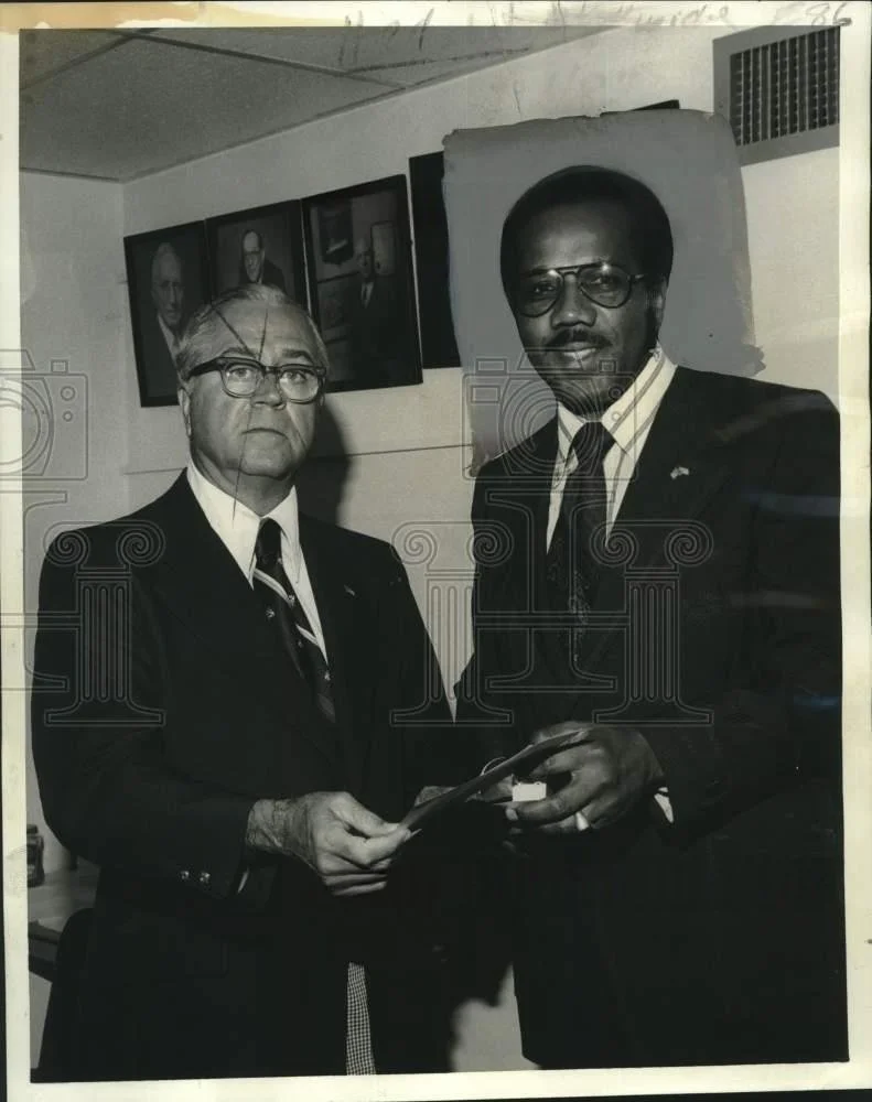 Two men in suits and glasses standing in front of a wall with framed photographs. They are holding a document or photograph together and looking at the camera.