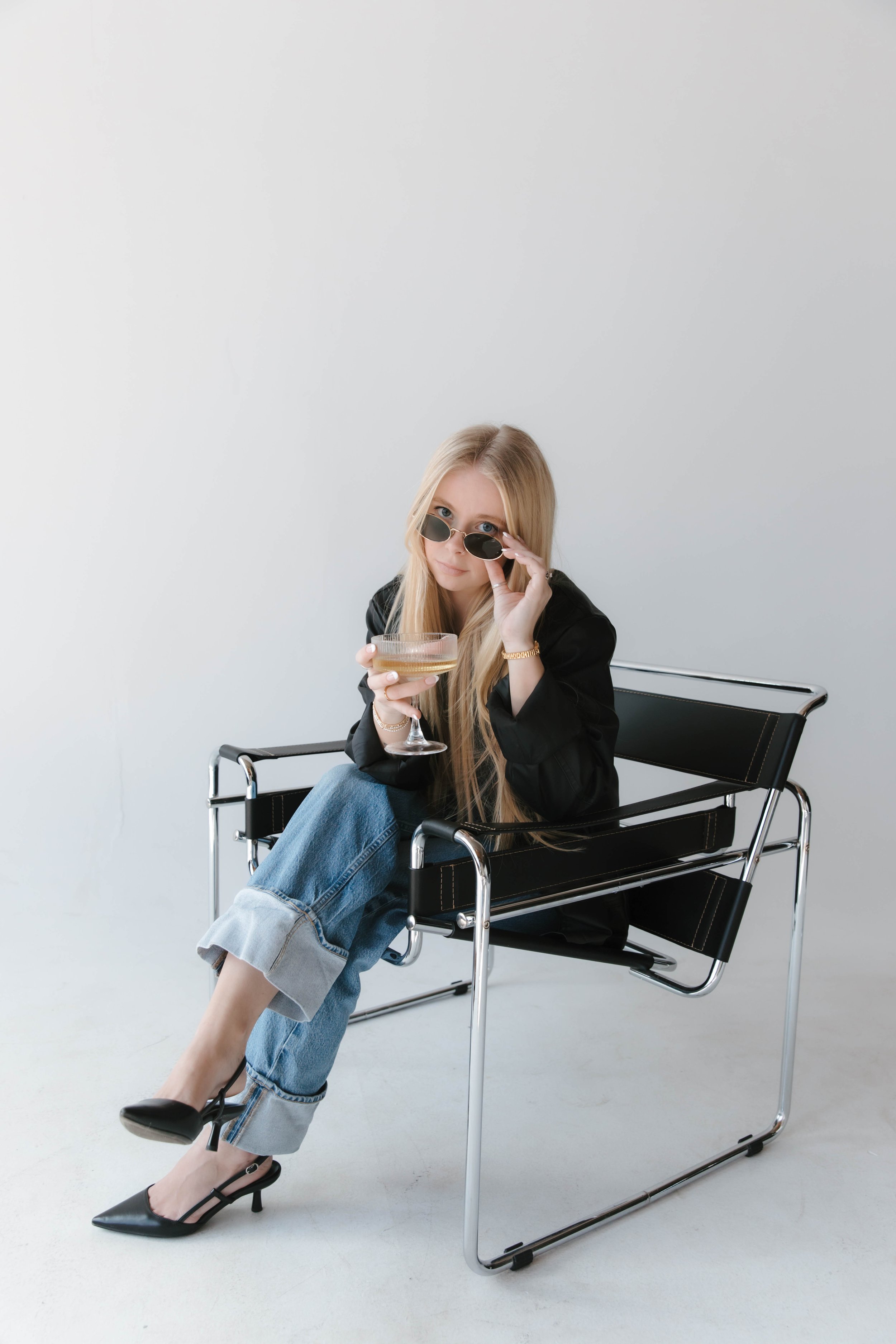 A woman with long blonde hair, wearing sunglasses, a black blazer, and light blue jeans with rolled cuffs, sitting on a black leather and chrome chair. She is holding a glass of champagne in her left hand and adjusting her sunglasses with her right hand. She is dressed elegantly with black high heels and a gold watch, against a plain white background.