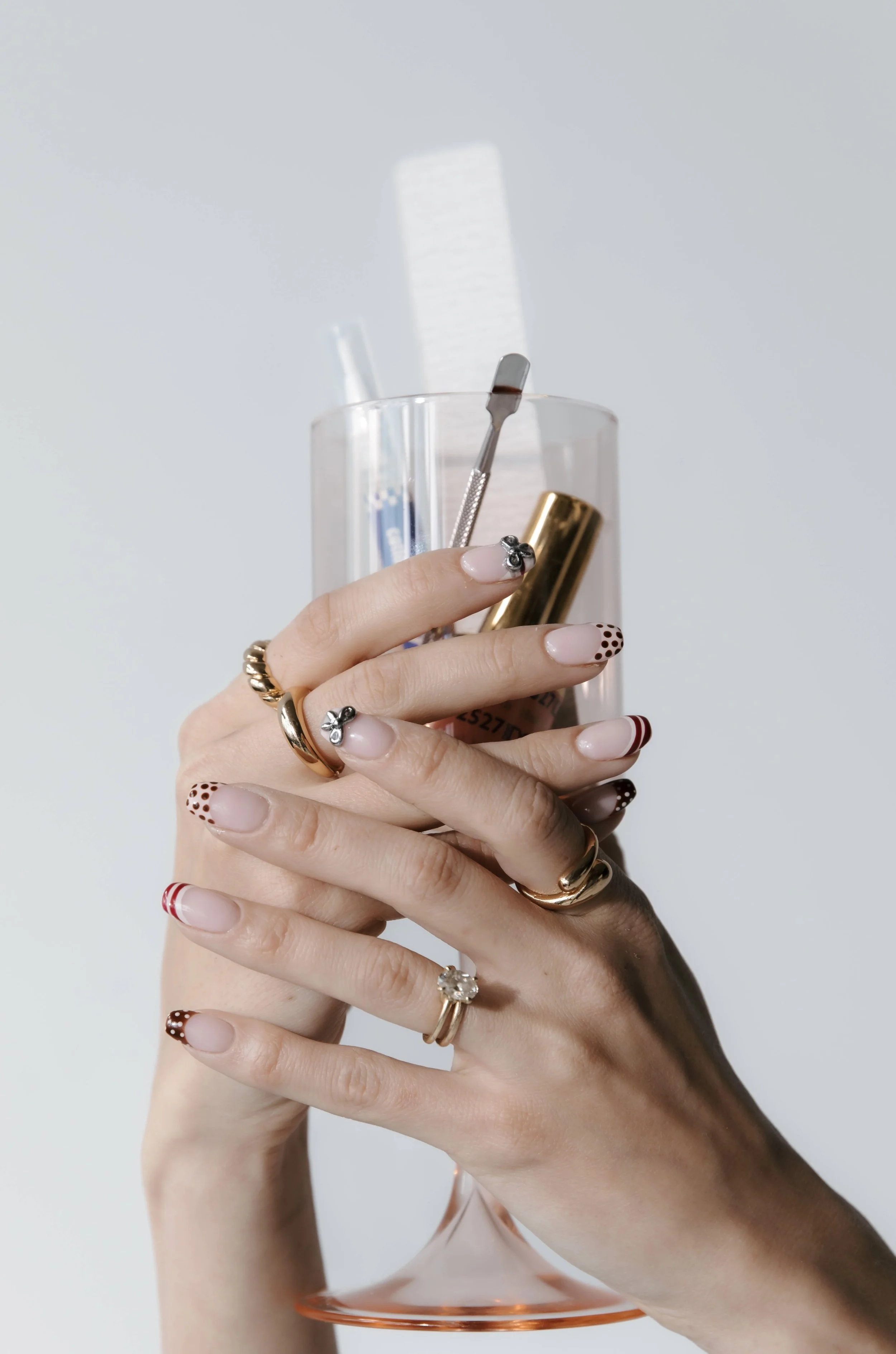 Close-up of a person's hands holding a glass container with nail polish tools, wearing rings and decorated nails, against a plain background.