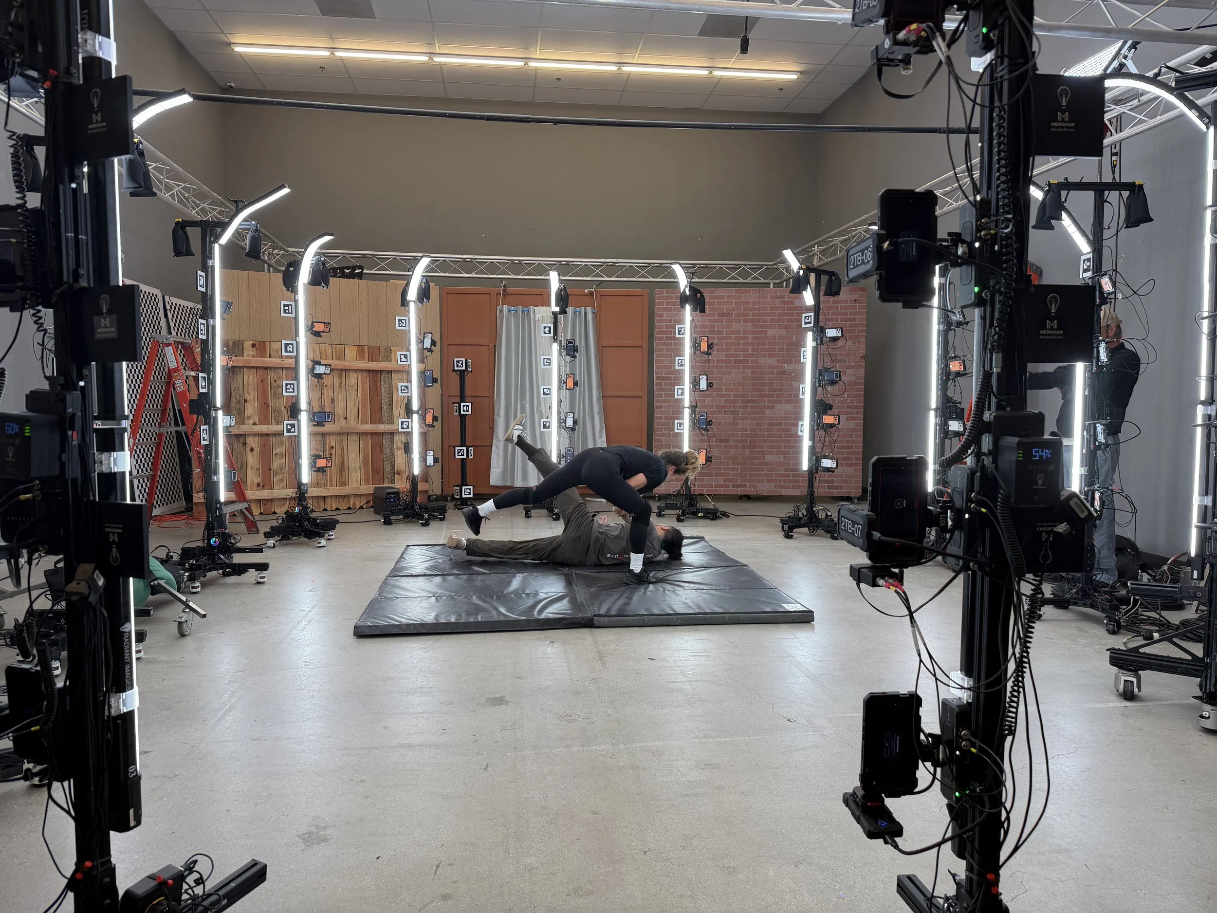 A photography or filming studio setup with a large circular rig equipped with multiple lights and cameras. Two people are on the floor, one person appears to be assisting or adjusting the other who is lying down on a mat. The background features a wa