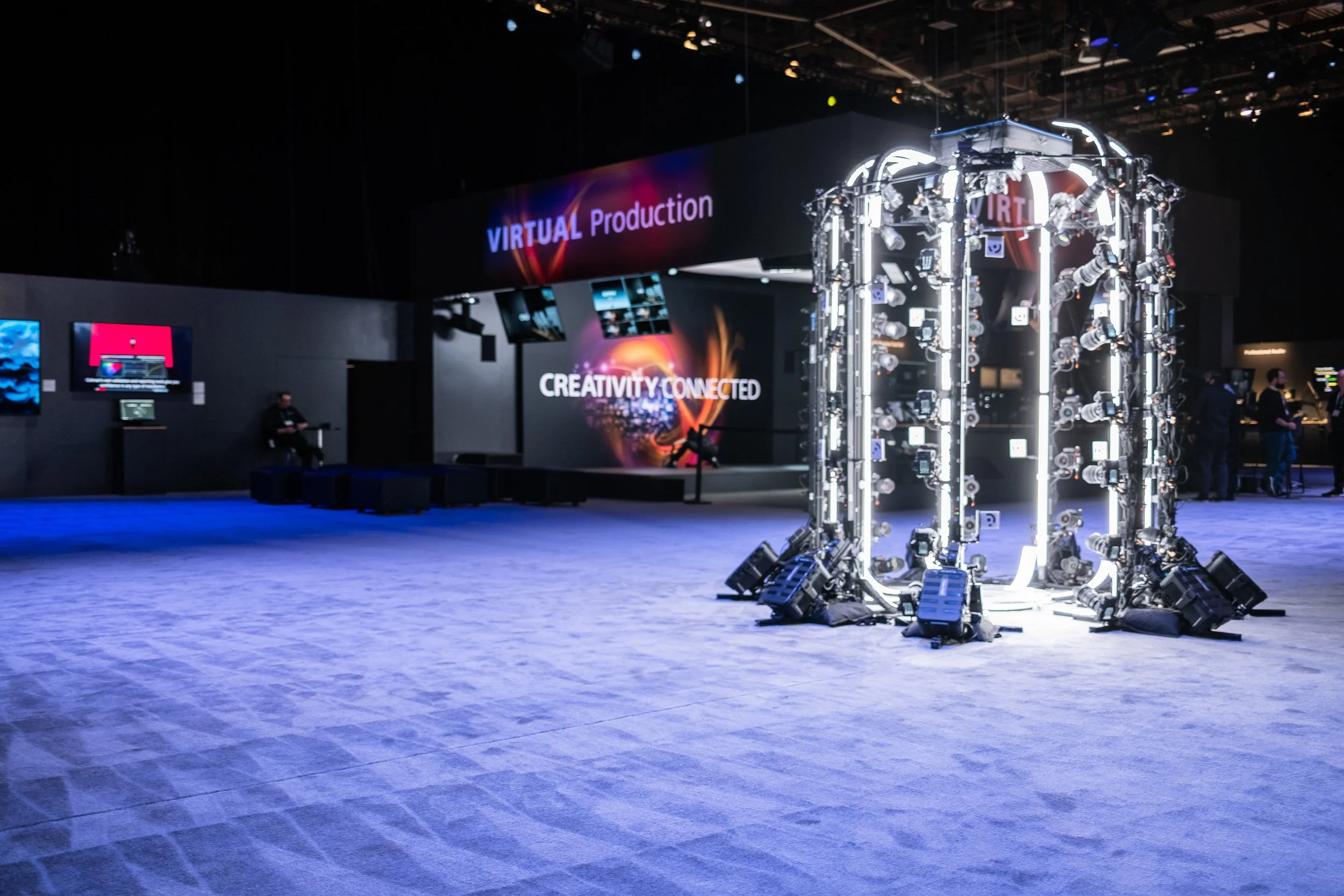 An exhibition space showing a large digital screen with the words 'VIRTUAL Production' and 'CREATIVITY CONNECTED'. There is a circular lighted structure made of numerous cameras or lights in the foreground and people walking or standing around in the