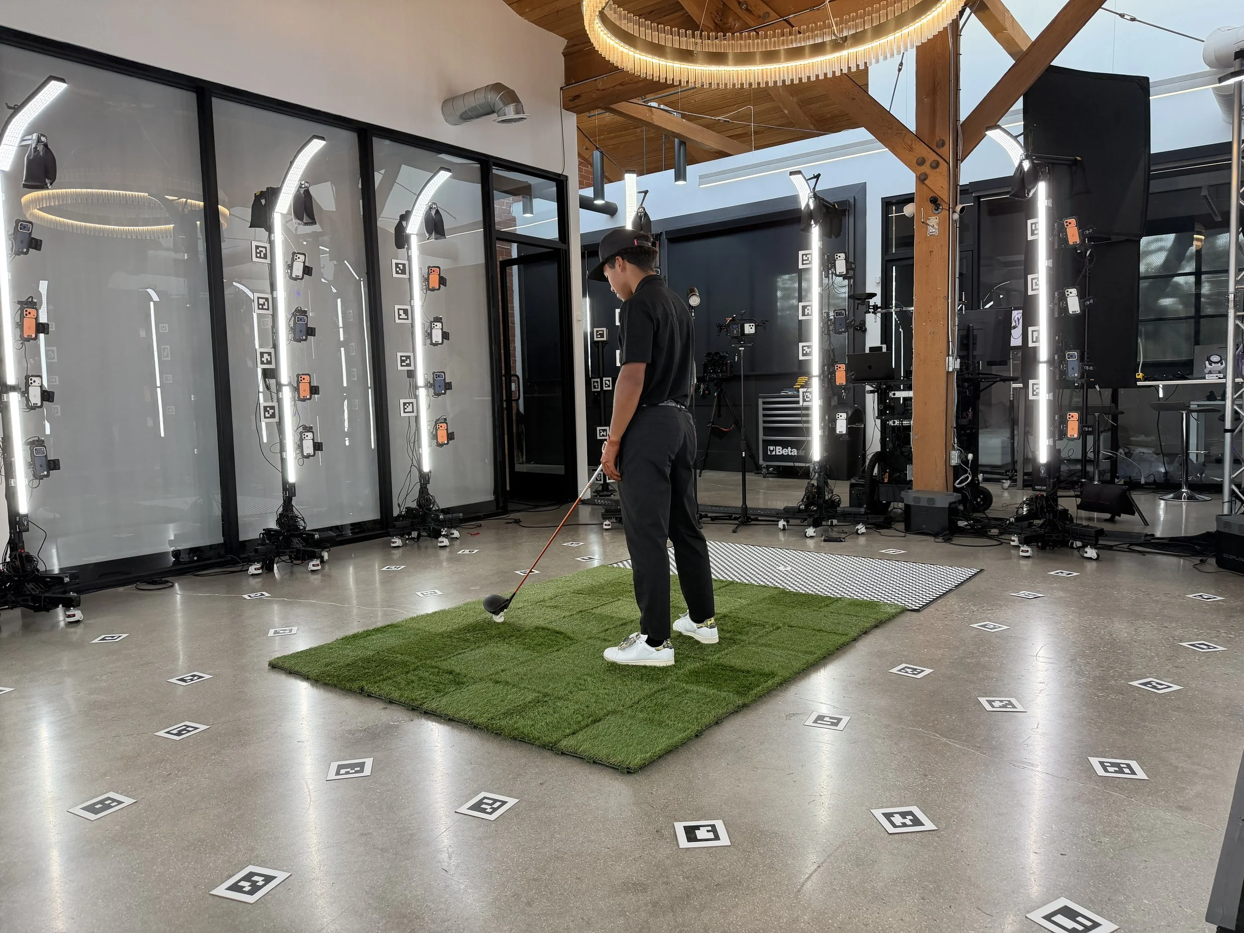 A person playing indoor golf with a golf club on a small green carpet, surrounded by a tech setup with cameras and lights in a room with industrial design. Meridian 4D portable volumetric gaussian splat system