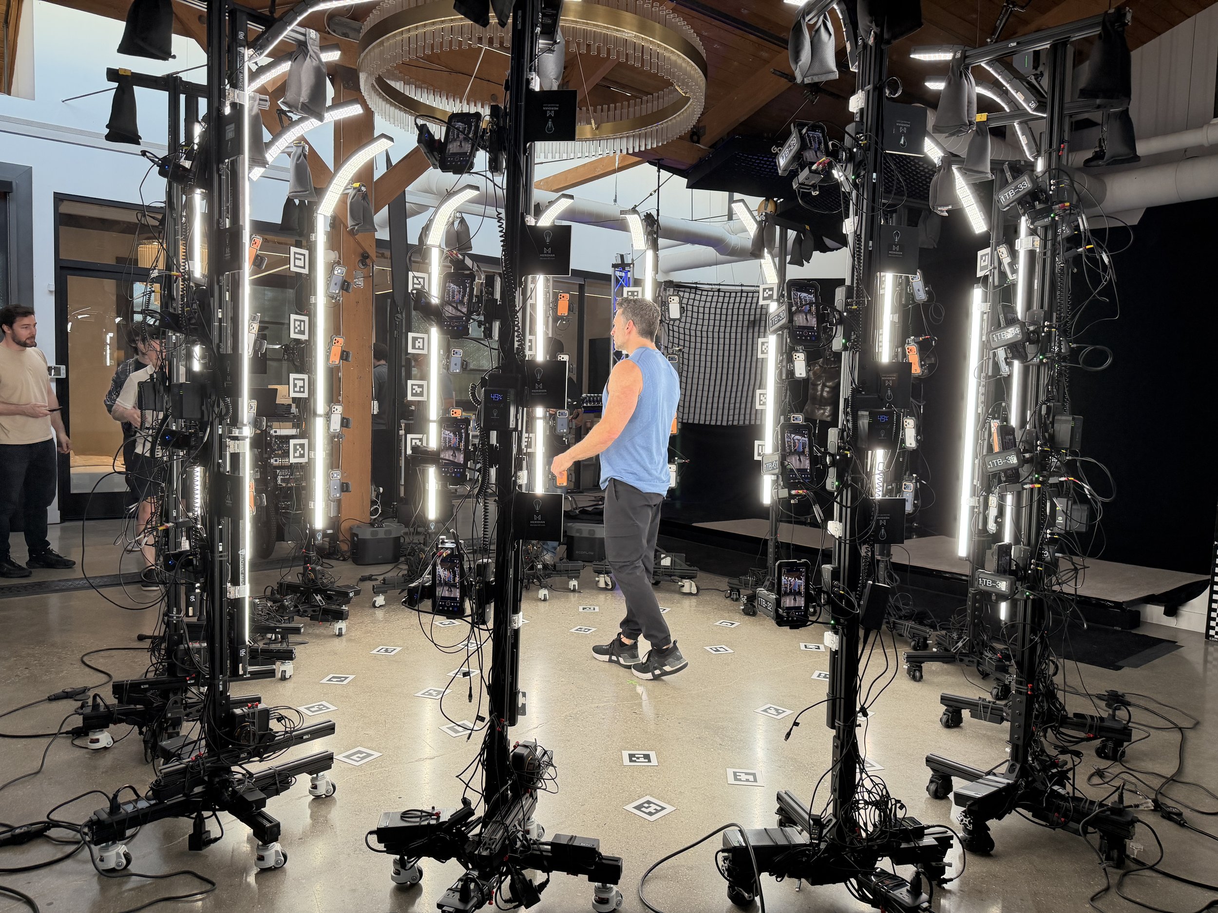 A studio setup with multiple vertical light rings and numerous mobile phones mounted on stands. There are people in the background and a person in a blue shirt walking among the equipment. The space has a high wooden ceiling with visible beams and re