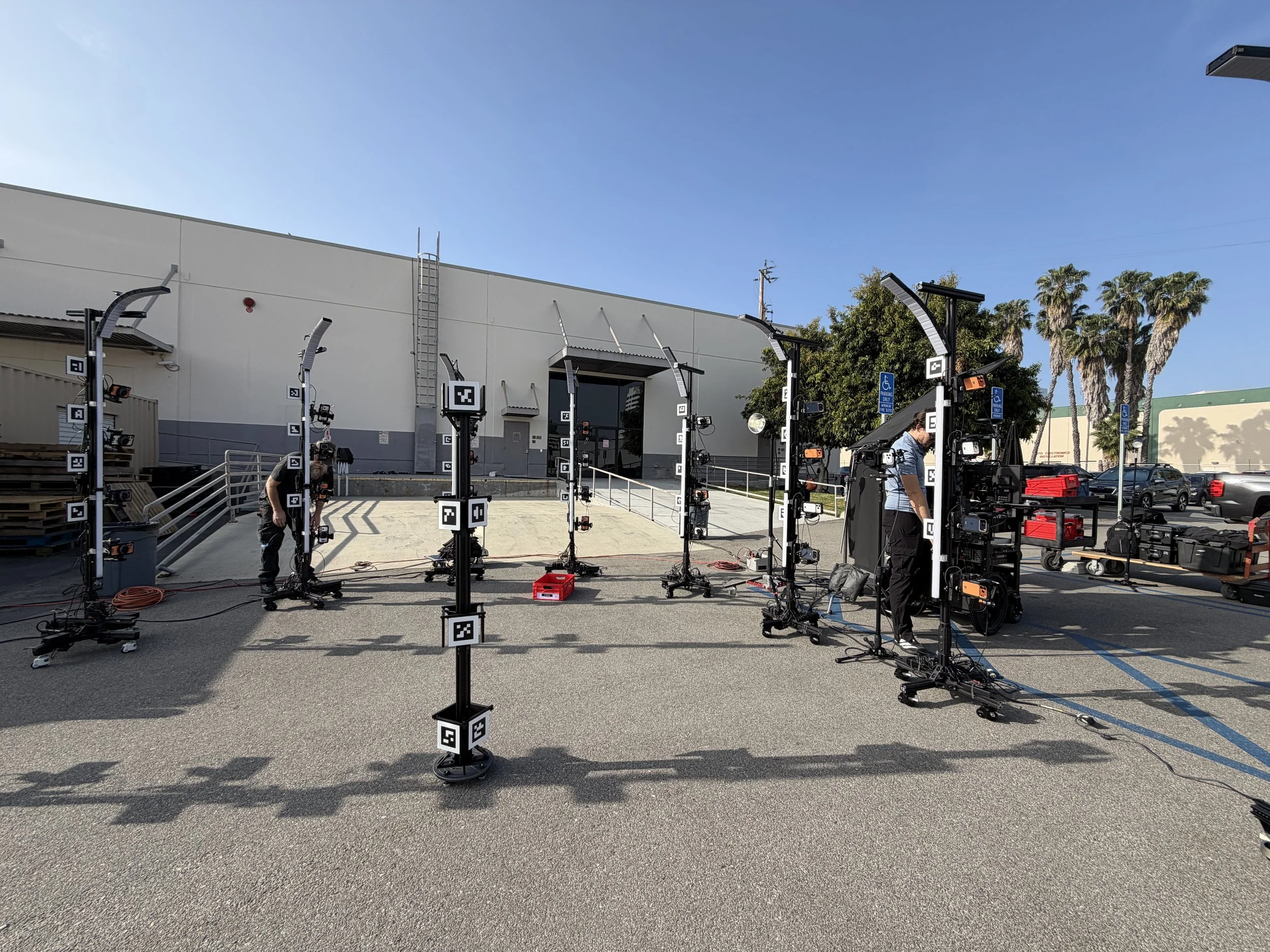 Outdoor scene with film equipment setup, including camera rigs, lights, and other gear in a parking lot near a building with palm trees and blue sky. Meridian 4D portable volumetric gaussian splat system