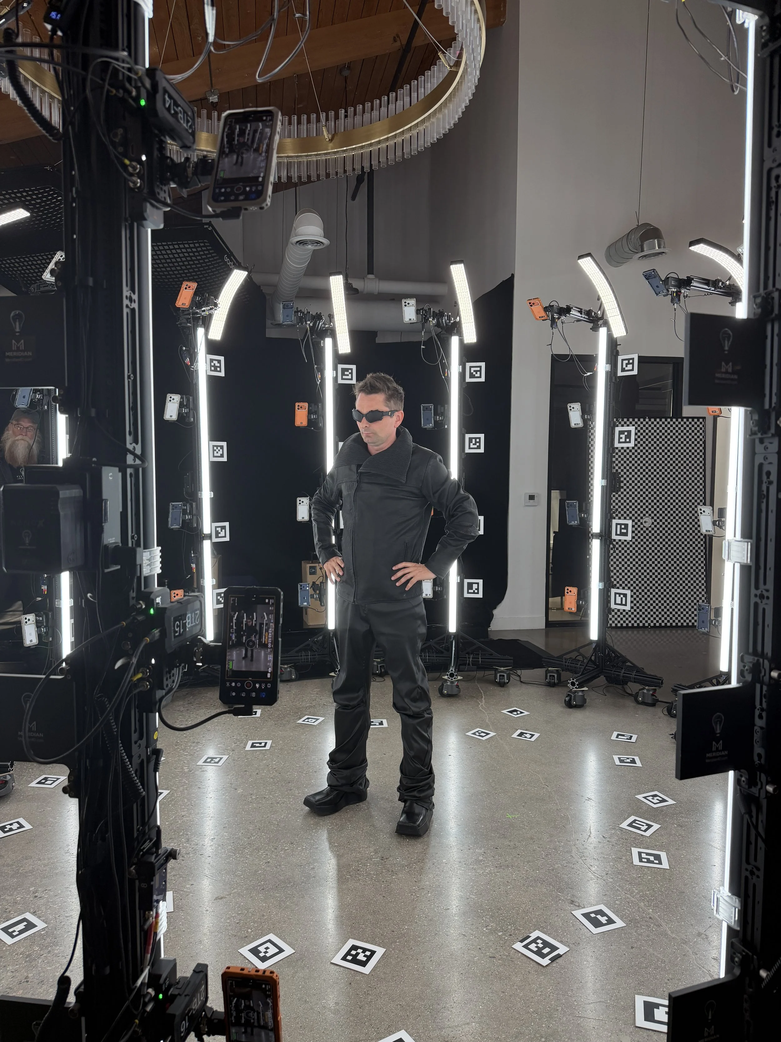 A man in black leather jacket and sunglasses posing for a photo in a studio with multiple LED lights and cameras. The studio has a modern design with a gray wall, a circular metallic chandelier, and floor tiles with QR code patterns.