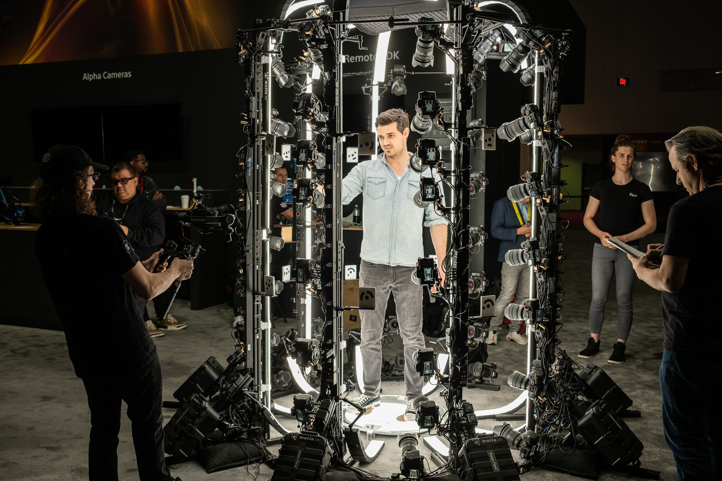 A man stands inside a circular array of cameras and lighting equipment, surrounded by crew members, during a video or photo shoot in a dark room.