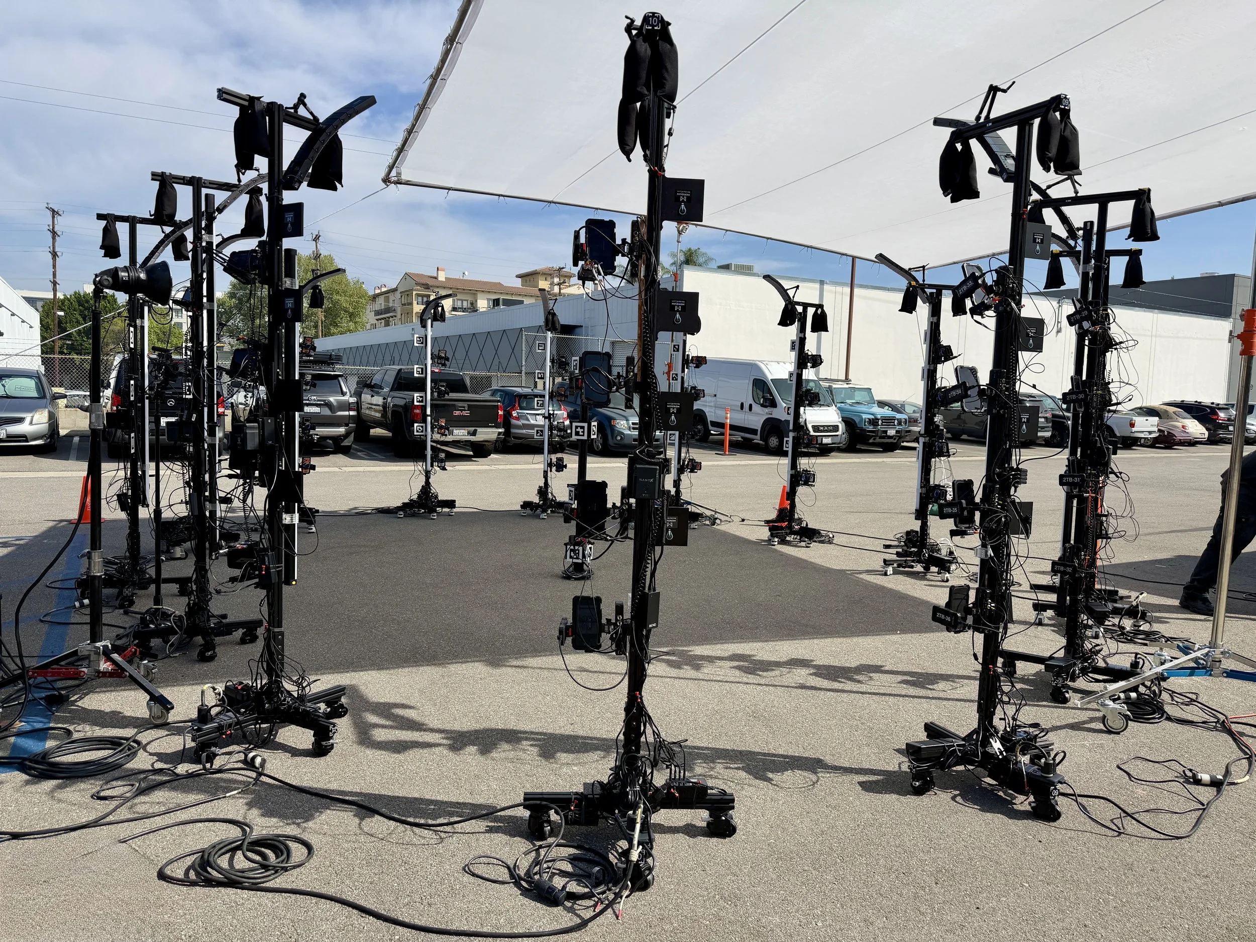 Outdoor setup with multiple camera equipment and lighting stands under a large white canopy in a parking lot, with cars and buildings in the background during daytime.