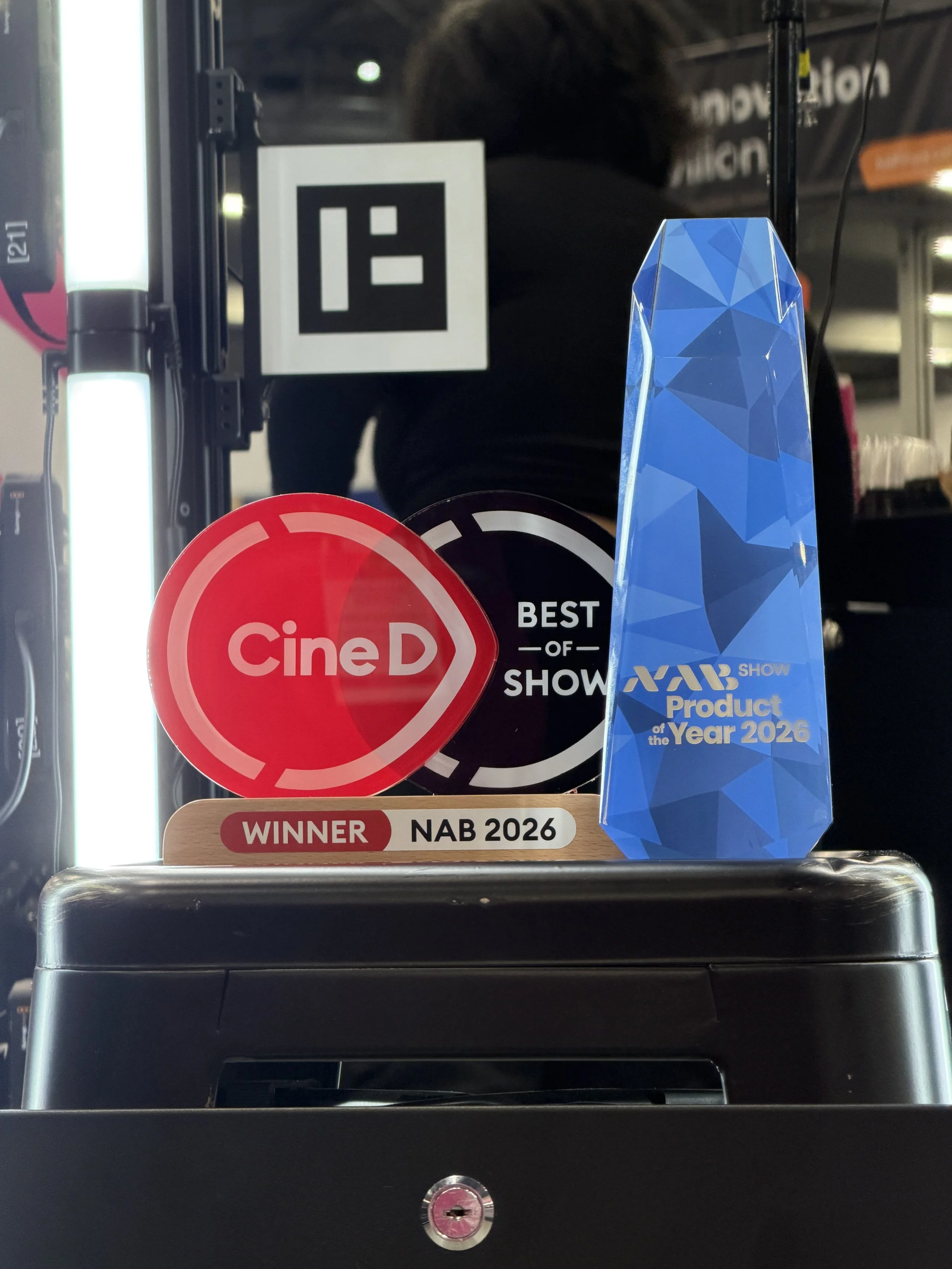 A display on a shopping cart shelf with awards and signs, including a red 'Cine D' sticker, a black 'Best of Show' sticker, a wooden plaque with 'Winner NAB 2026' written on it, and a blue geometric trophy labeled 'NAB Product of the Year 2026.'