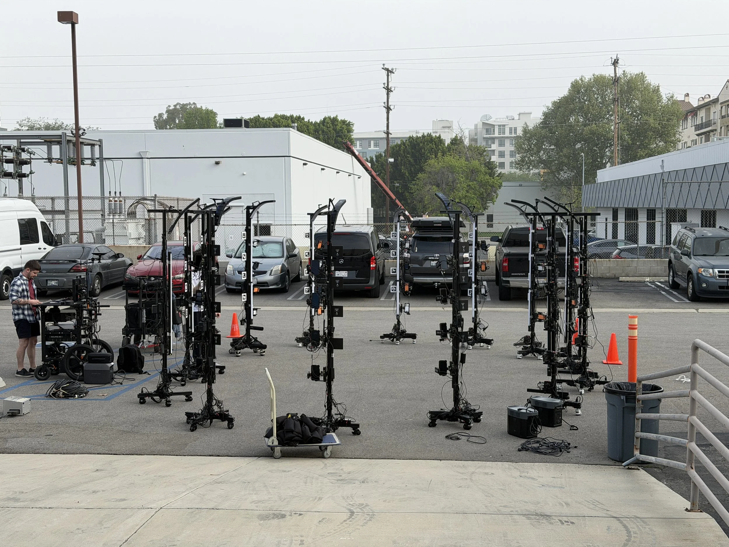 Setup of multiple film or photography lighting equipment in a parking lot with a person operating one of the devices, background includes parked cars, industrial building, and residential buildings.