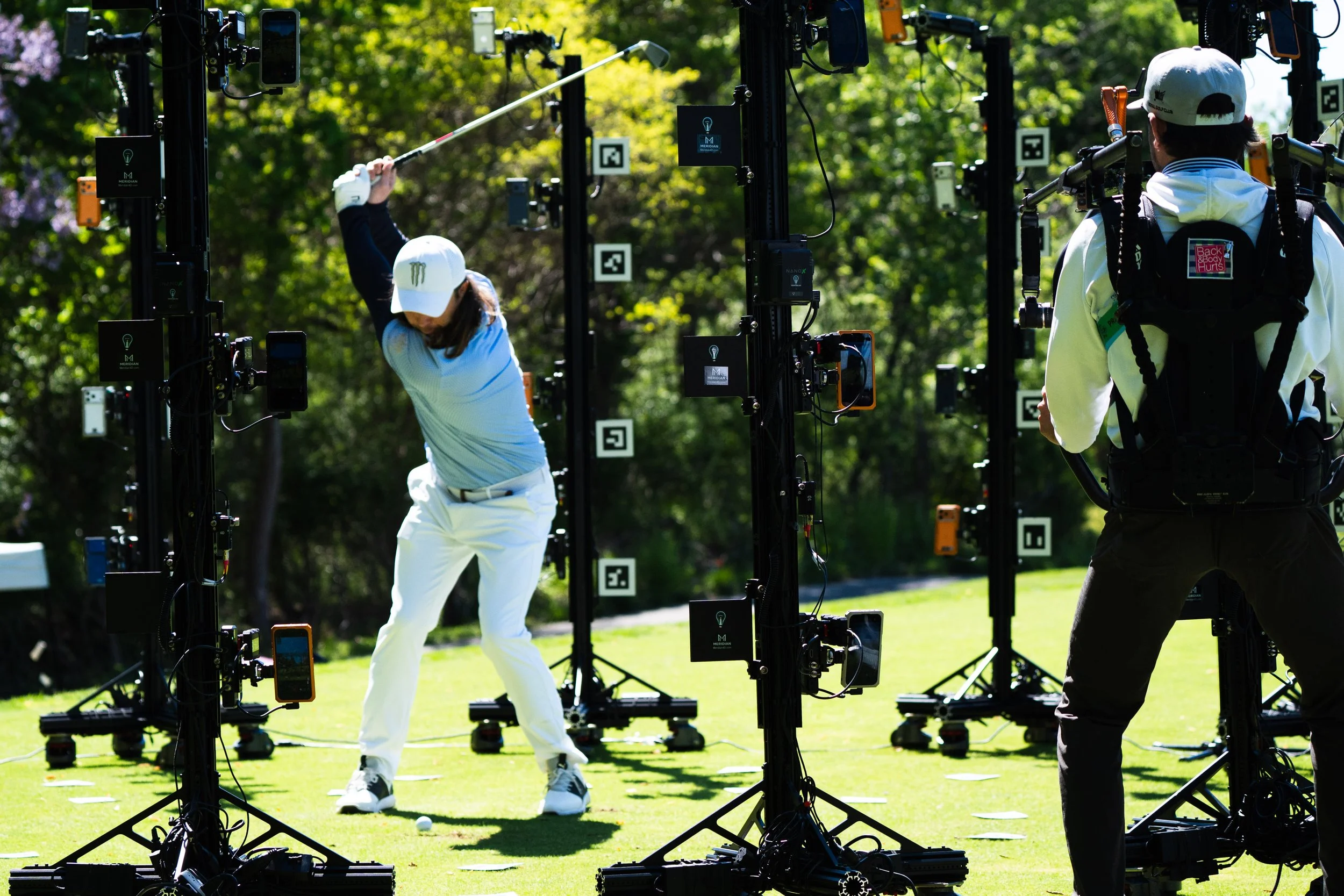 A person practicing golf swings at an outdoor driving range with a golf club, surrounded by technology equipment and a coach or instructor observing, under a sunny sky with trees in the background.