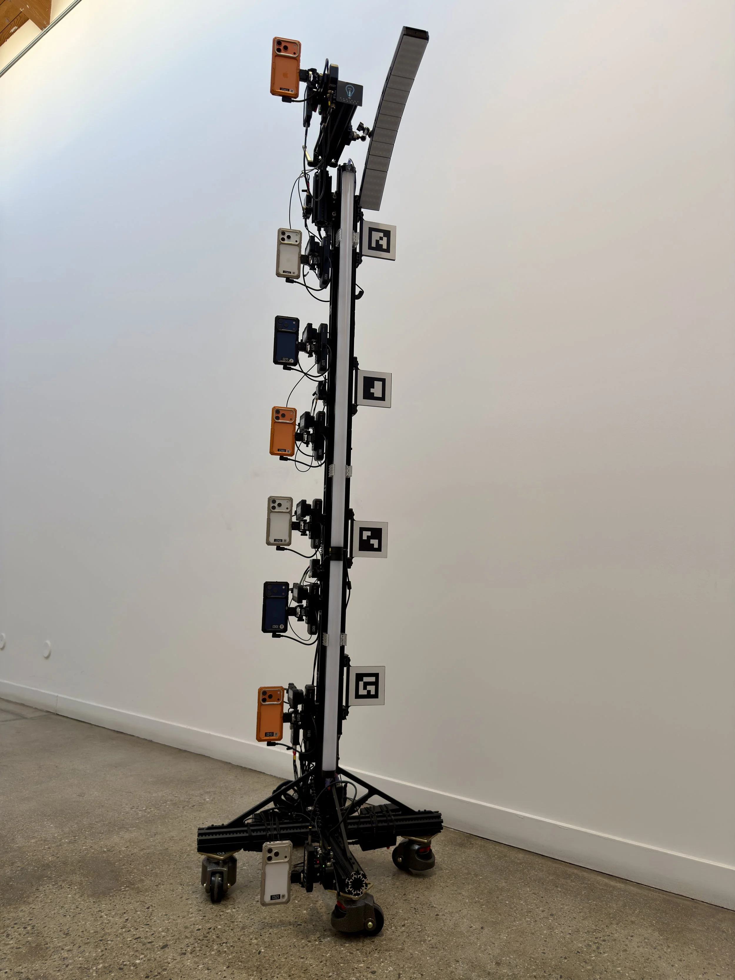 A tall robotics setup with multiple cameras and sensors on a black stand, placed indoors against a white wall, with mounted electronic devices and cables on a black wheeled base. Meridian 4D portable volumetric gaussian splat system