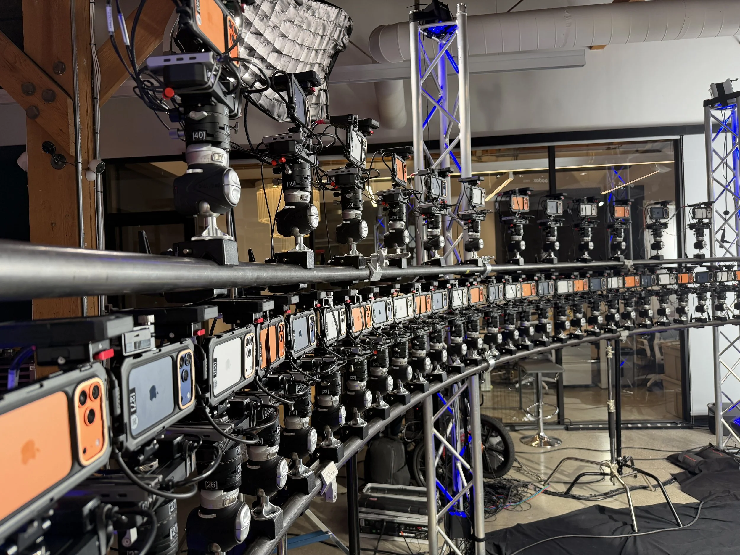 A setup of multiple iPhones mounted on robotic rigs on a metal frame, with some electronic equipment and cables on the floor, likely used for testing or recording.