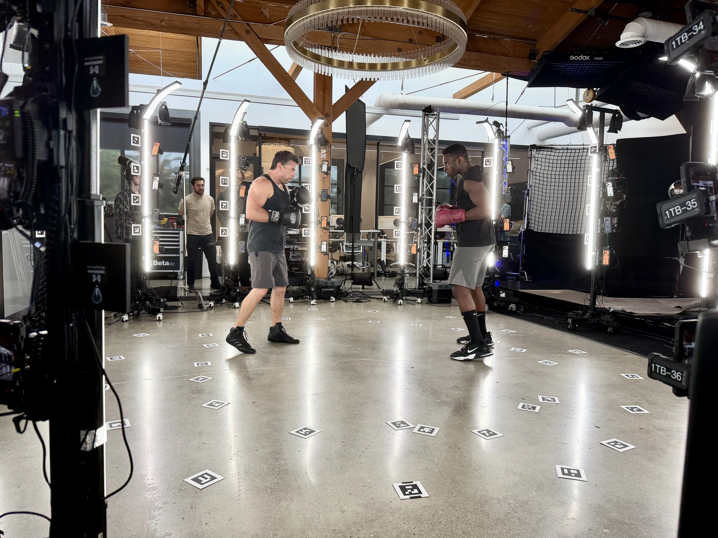 Two boxers are practicing with gloves in a gym, standing face to face underneath a large circular light fixture, with cameras and equipment around them. captured in volumetric video and Gaussian splatting 
