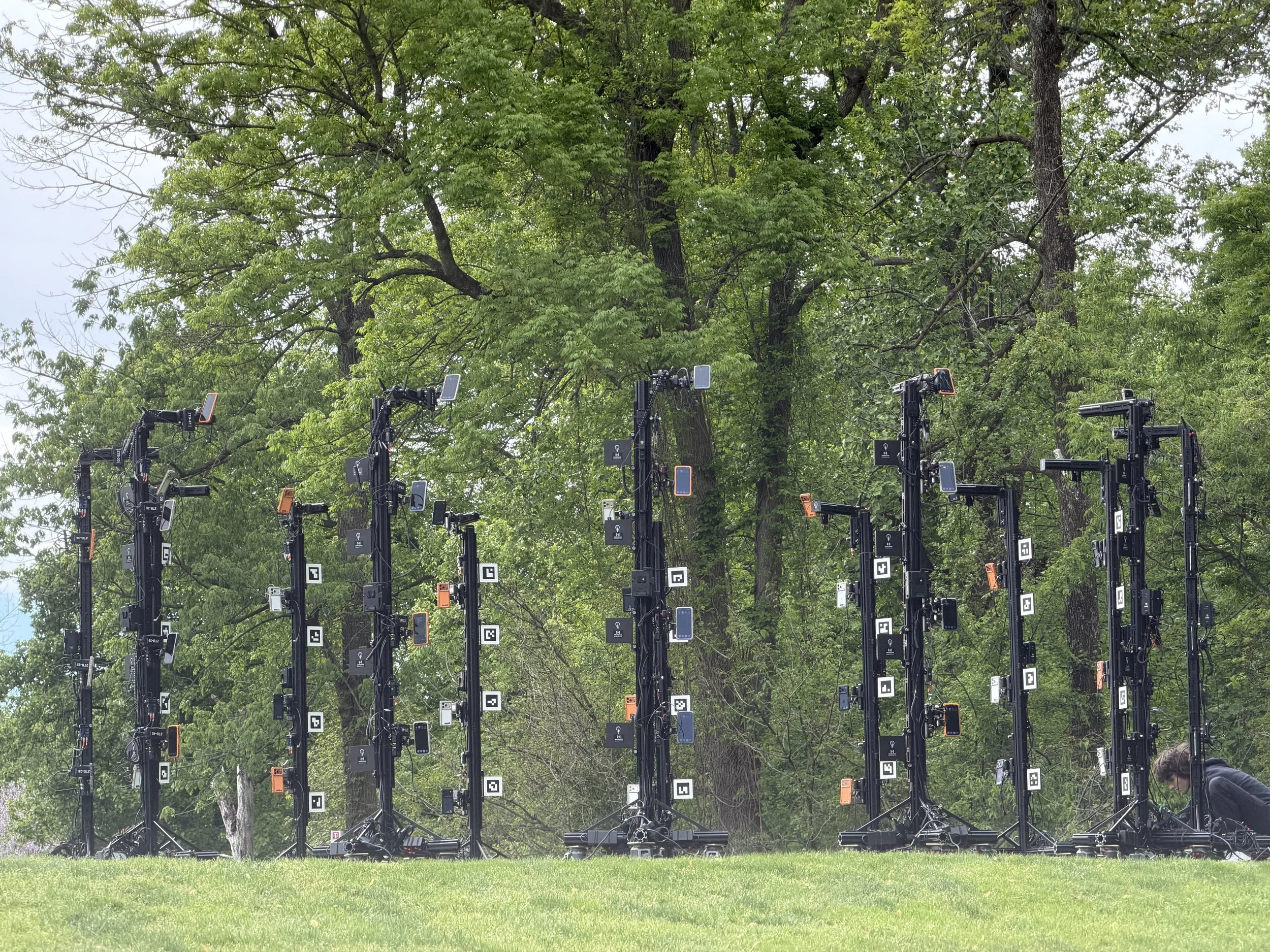 A row of camera tech equipment set up outdoors in front of green trees, with a person in the bottom right corner.