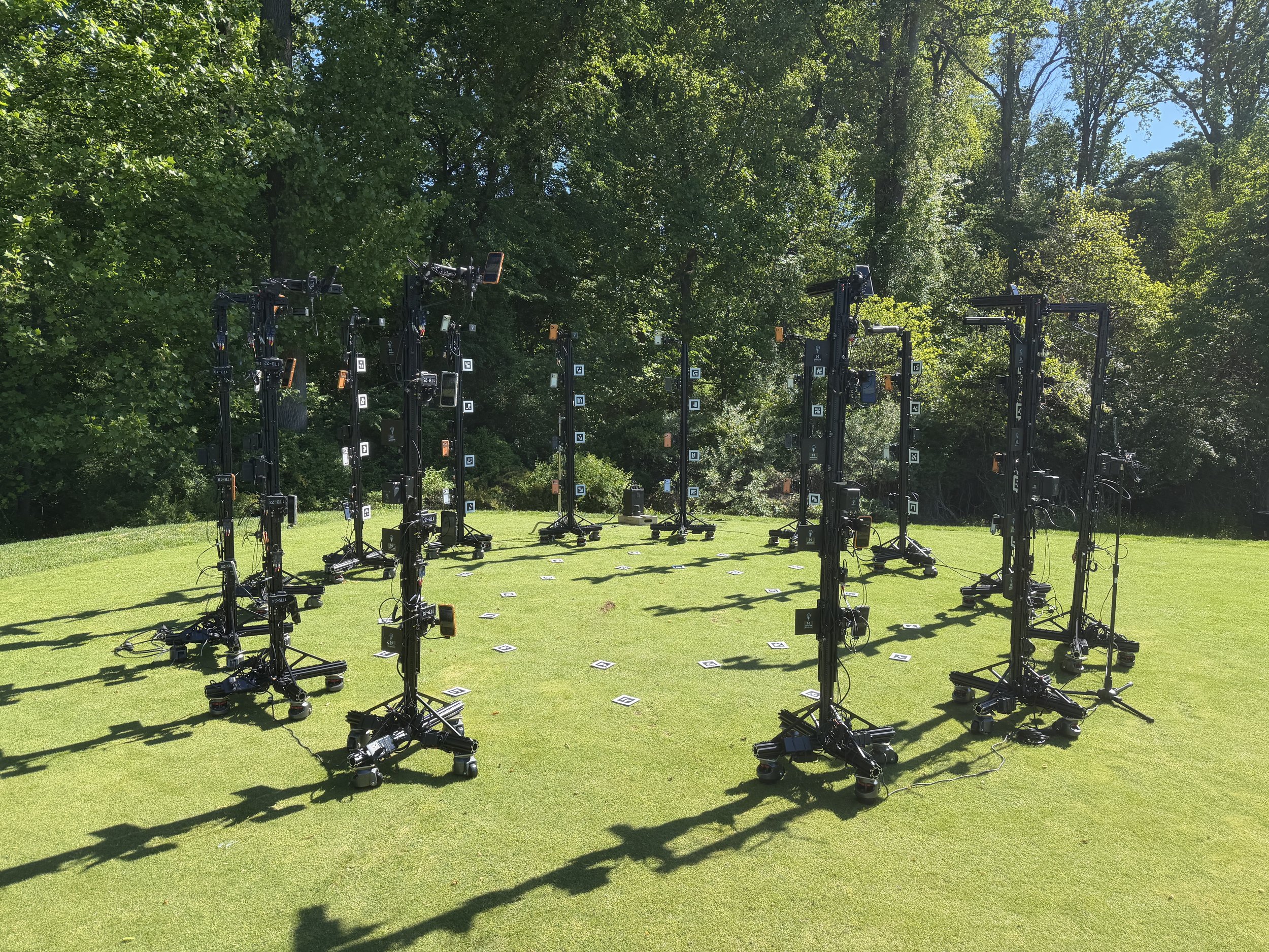 A setup of numerous black camera equipment on tripods arranged on a grassy lawn in a park area with tall green trees in the background under a partly cloudy sky.