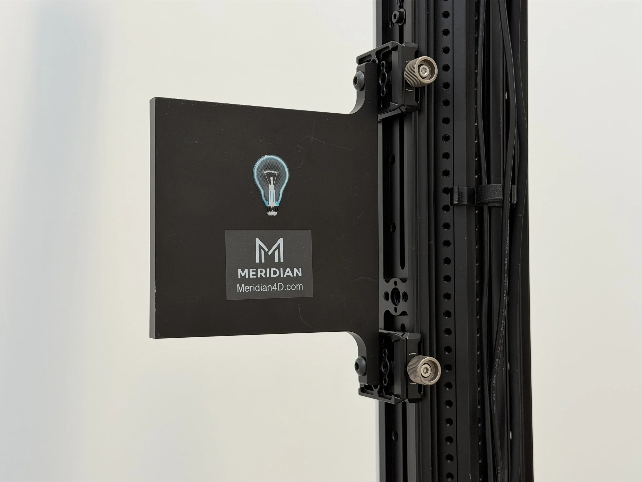 Close-up of a black metal structure with threaded knobs and adjustable components, featuring a loyalty sticker with a light bulb icon and the branding 'MERIDIAN' and website 'Meridian4D.com' on a white background.