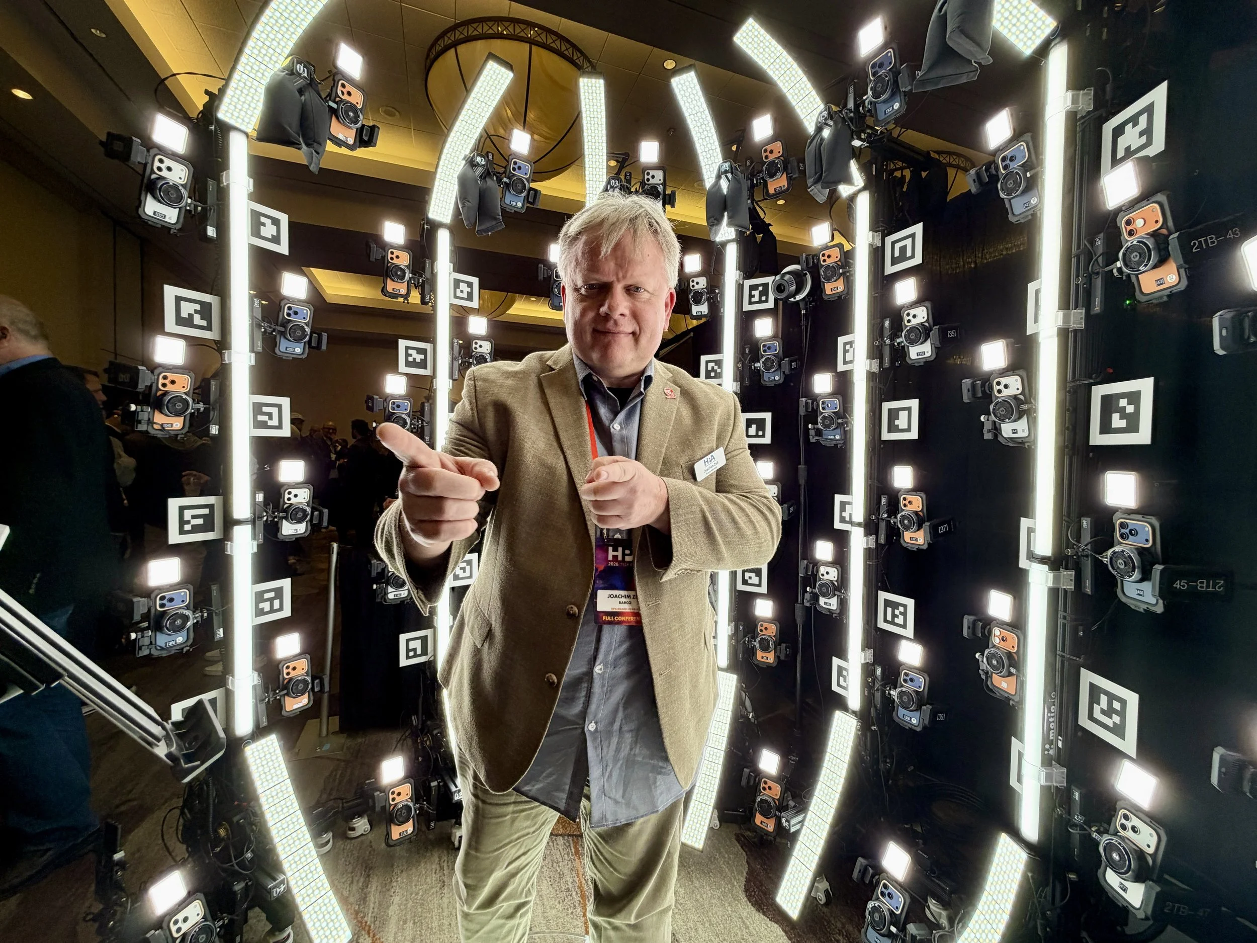 A man with gray hair and a beige blazer points towards the camera in front of a display of numerous cameras and lights arranged vertically and horizontally in a circle. HPA Meridian 4D portable volumetric gaussian splat system
