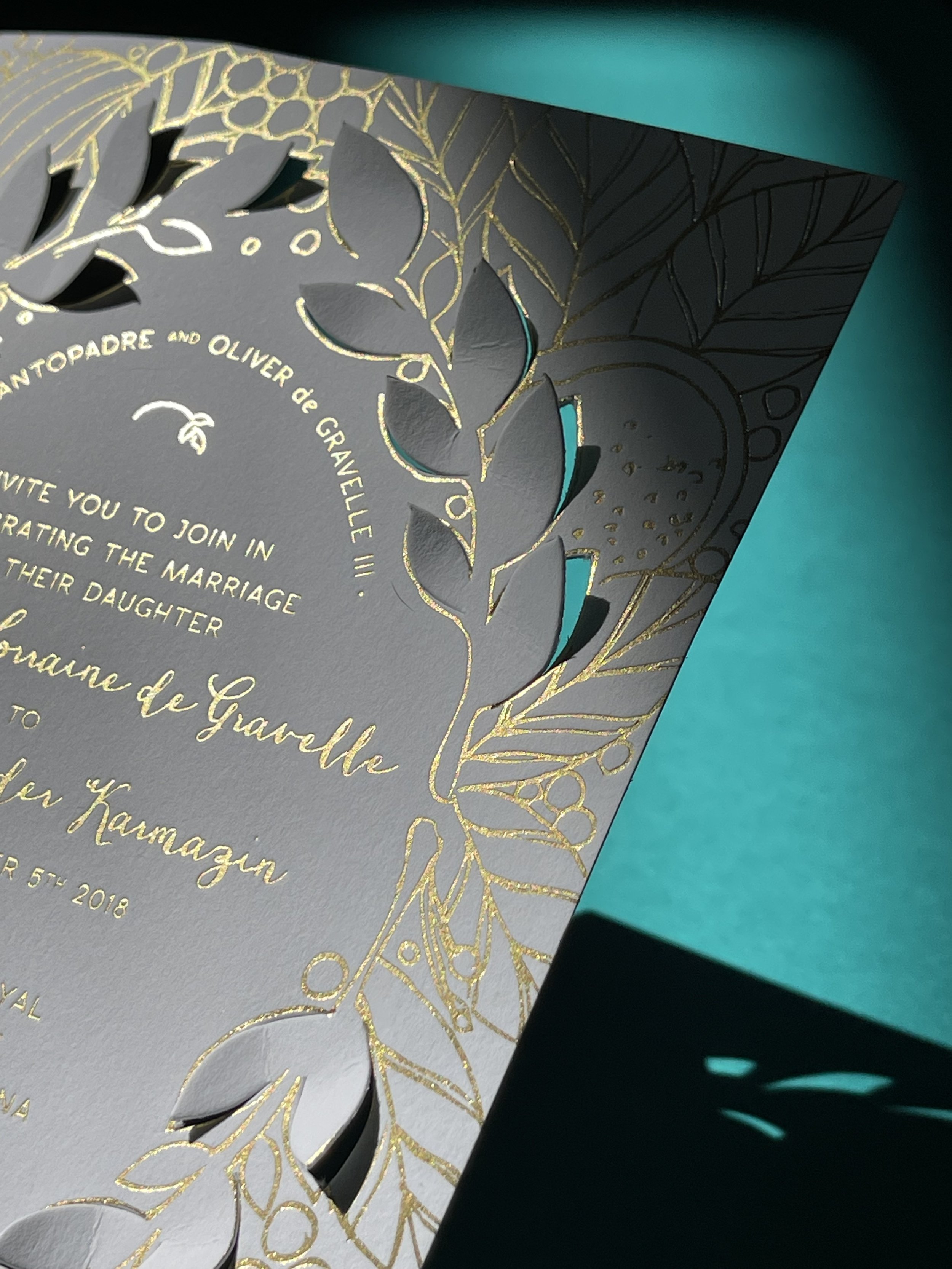Close-up of a wedding invitation with gold foil accents and cut-out design, featuring botanical patterns and cursive script, set against a teal background.