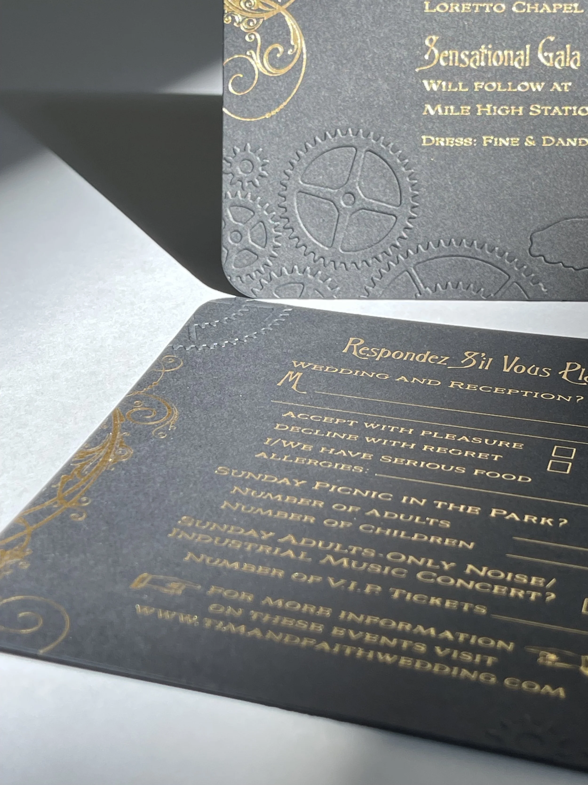 A dark-colored wedding invitation with gold embossed text and decorative gears in the background. The invitation includes details about a wedding and reception at Loretto Chapel, along with RSVP options and other event information.