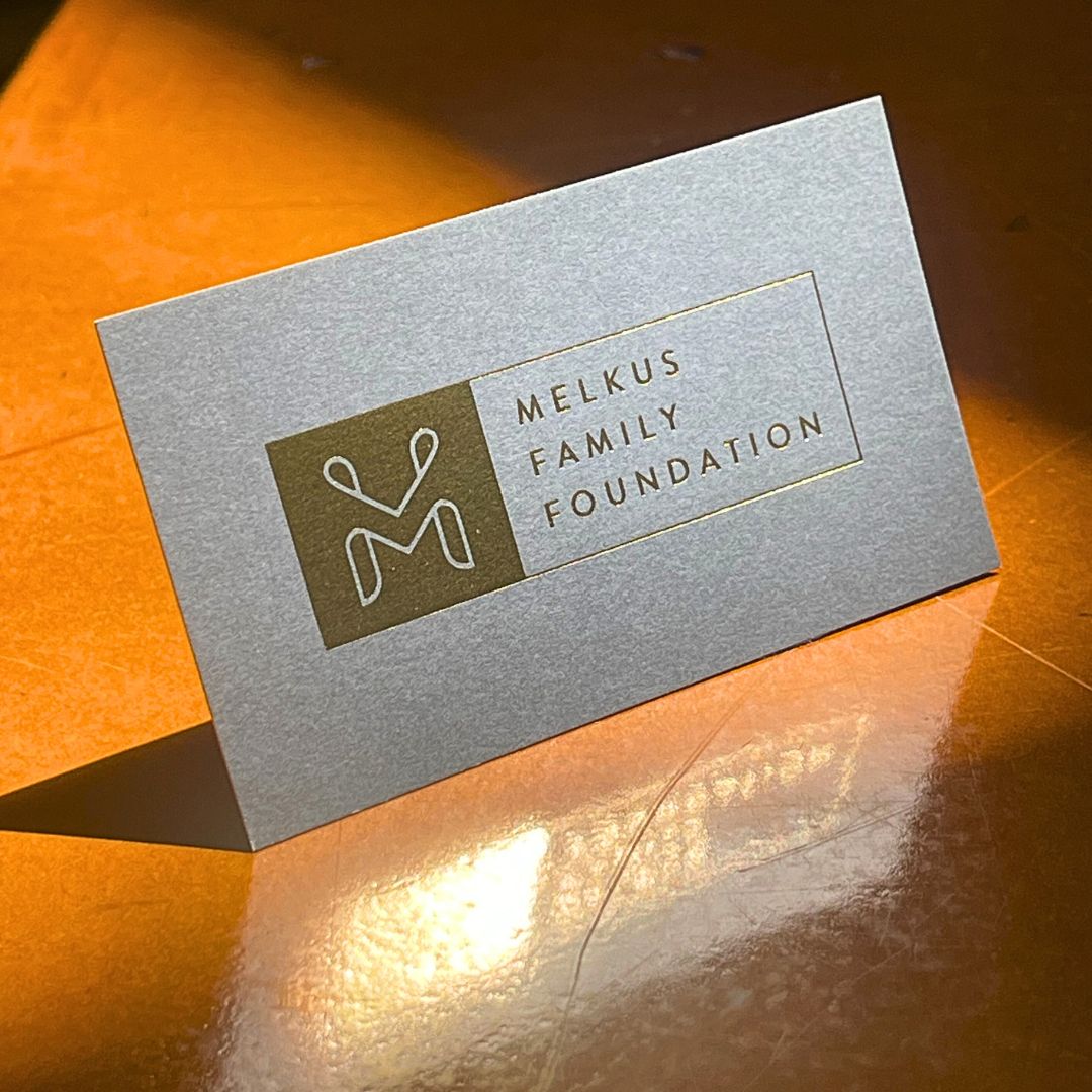 A silver reception card for the Melkus Family Foundation with a logo featuring a stylized figure, placed on a wooden surface with a warm light shining on it.