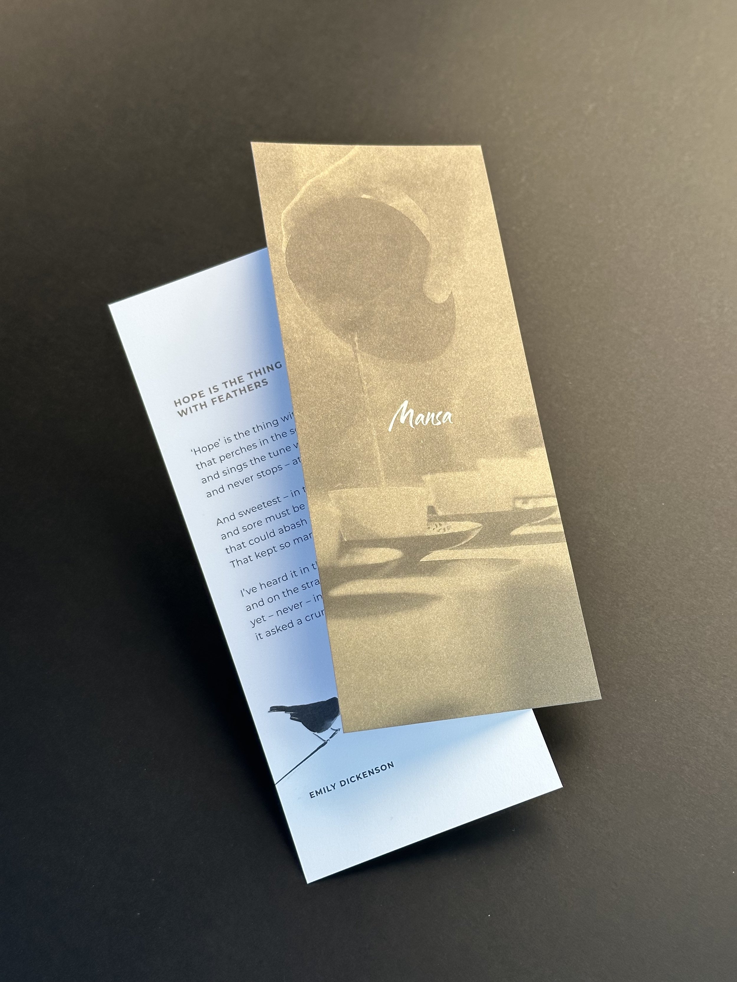 Close-up of two overlapping bookmarks, one with gold and the other with white background. The white bookmark contains poetic text, and the gold bookmark has an abstract bird silhouette and the word 'Mansa' written on it.