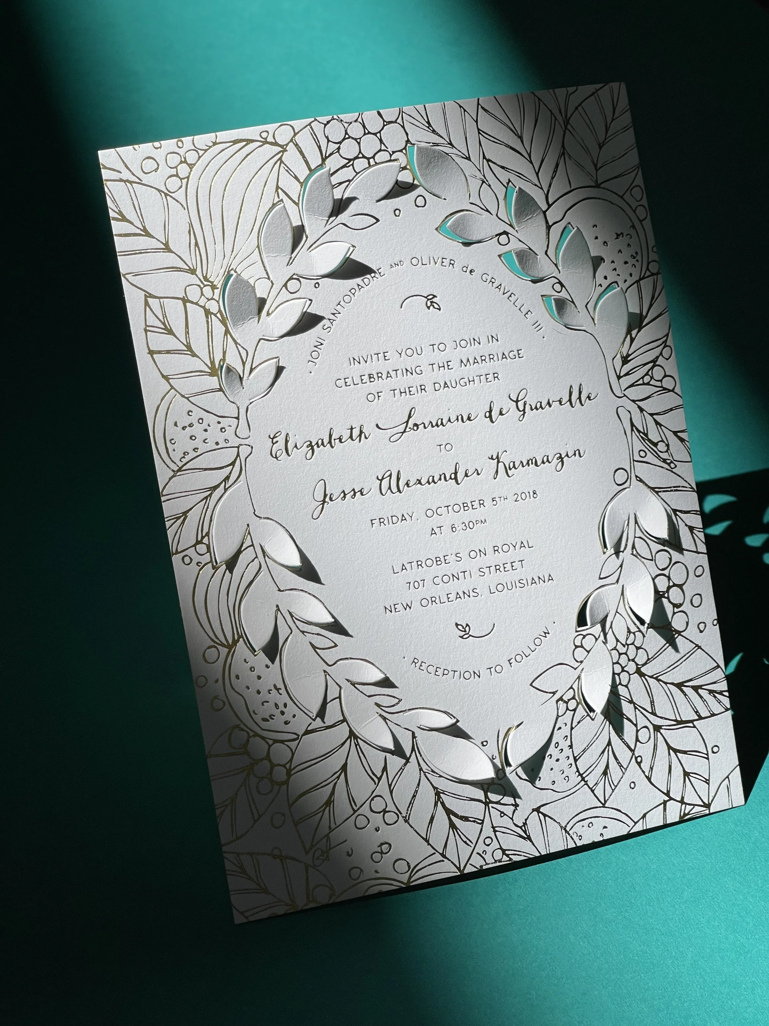 Wedding invitation with a floral gold foil design and cutout details on white paper, inviting guests to a celebration in New Orleans.