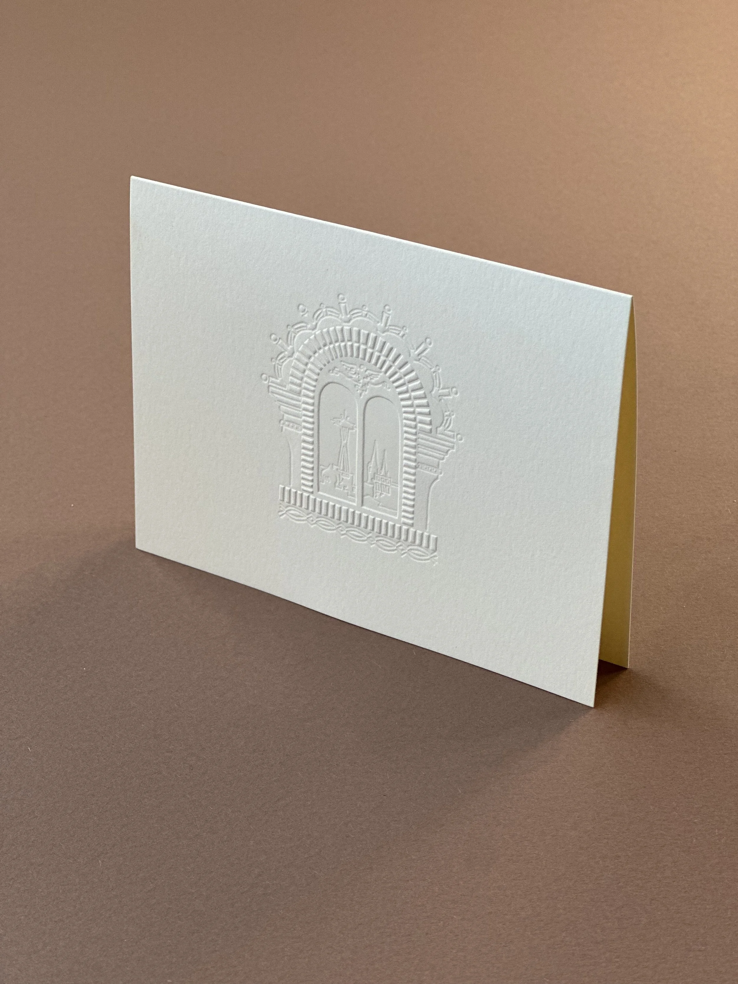 A folded white greeting card with an embossed religious window design, standing on a brown surface with a beige background.