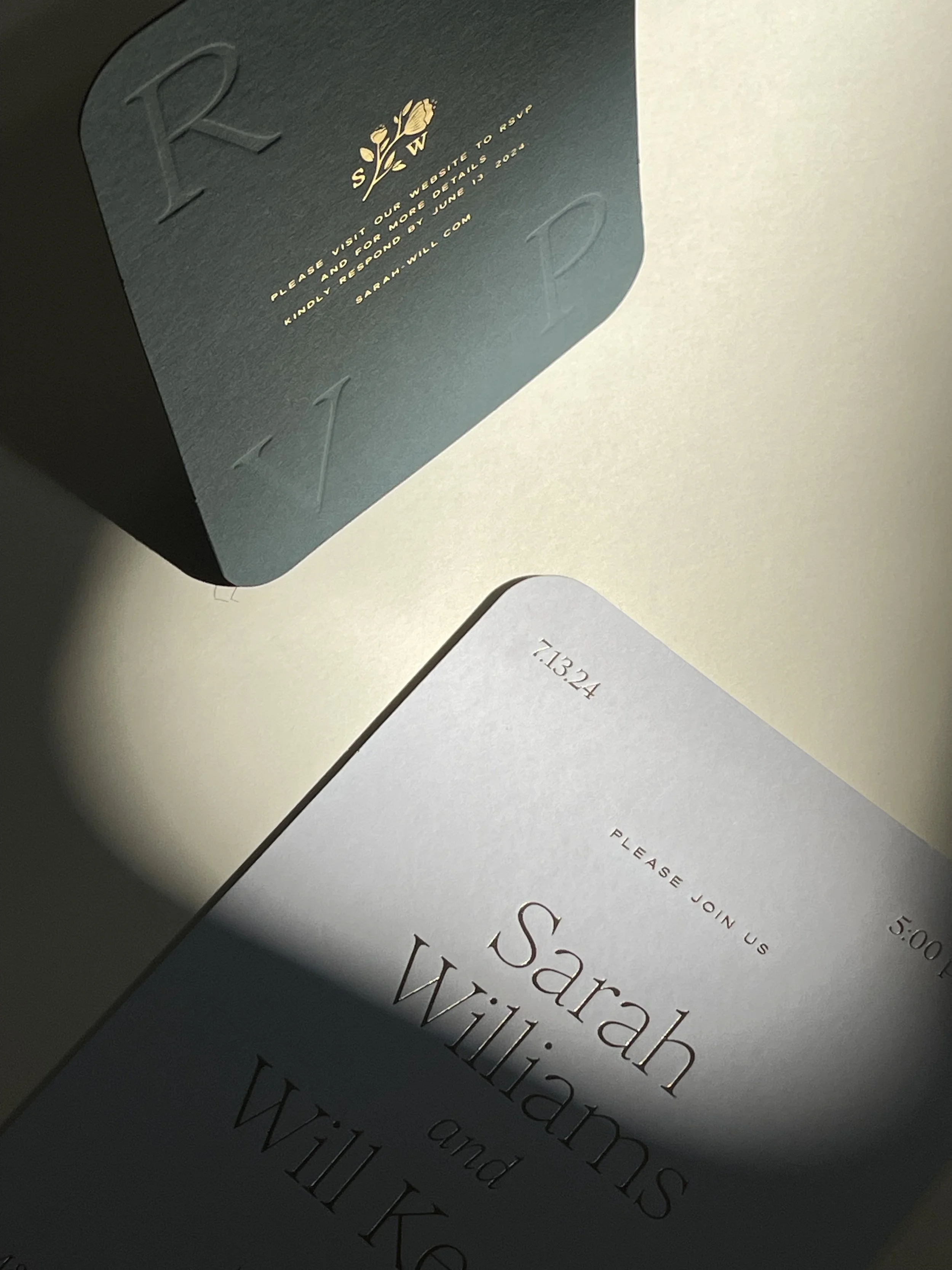 Wedding invitation cards with elegant text, one card in dark color with embossed letters and a gold floral emblem, the other card in light color with details of the wedding of Sarah Williams and Will Kess, including the date and time.