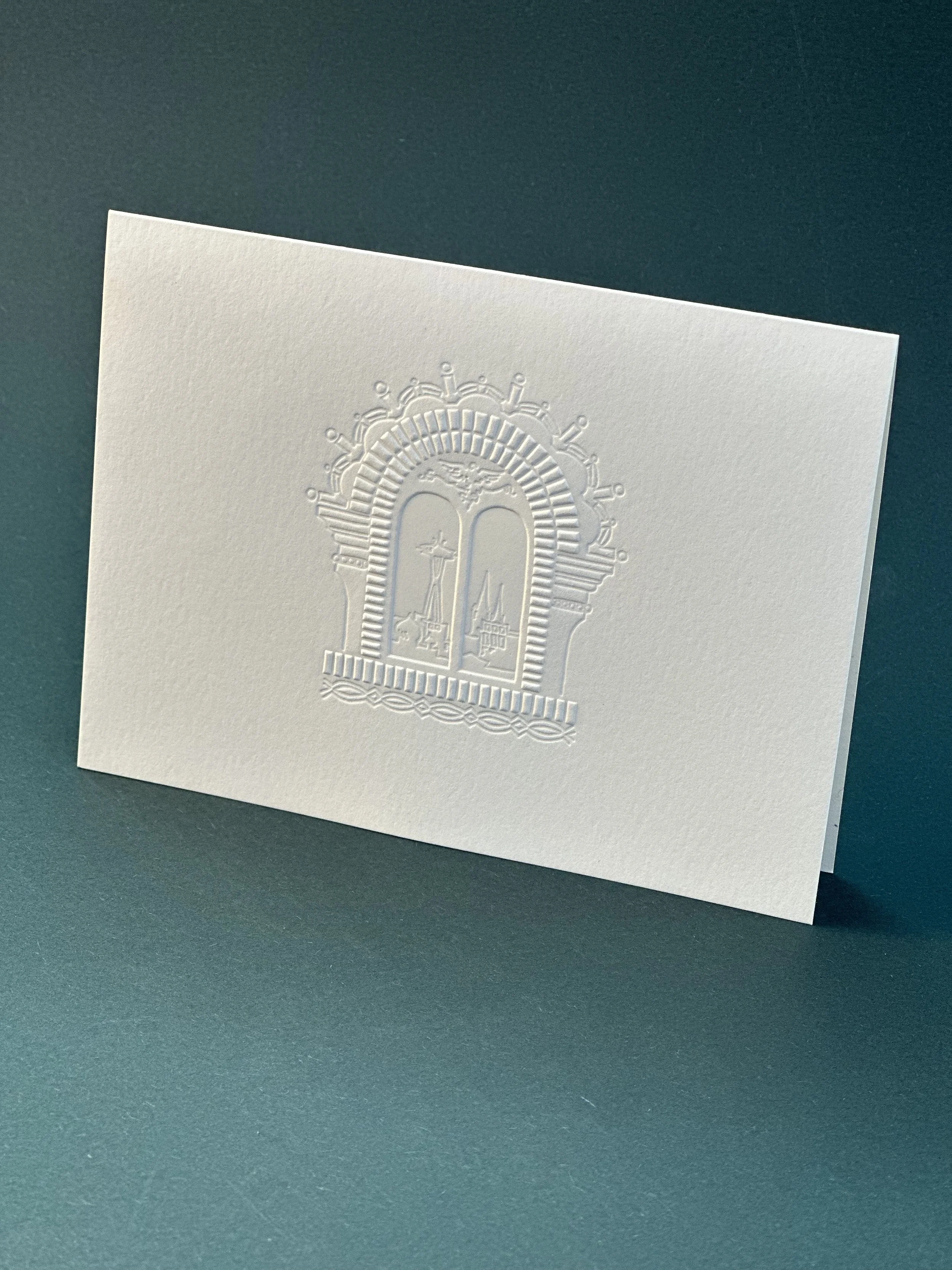White embossed greeting card featuring an intricate window arch design with multiple towers and architectural details, set against a teal background.
