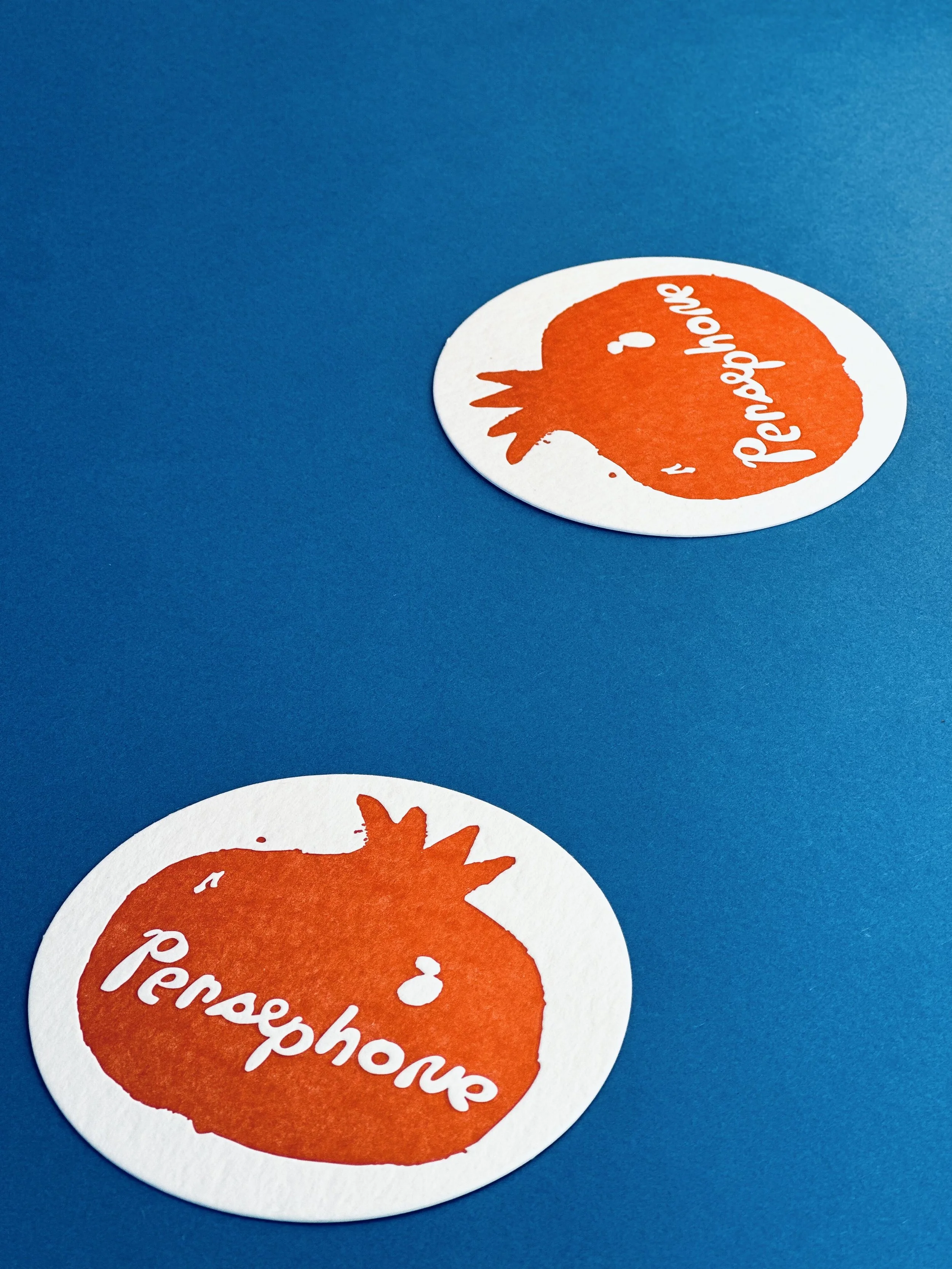 Two round stickers on a blue surface, each featuring an orange pomegranate illustration with the word 'Pomegranate' written across it.
