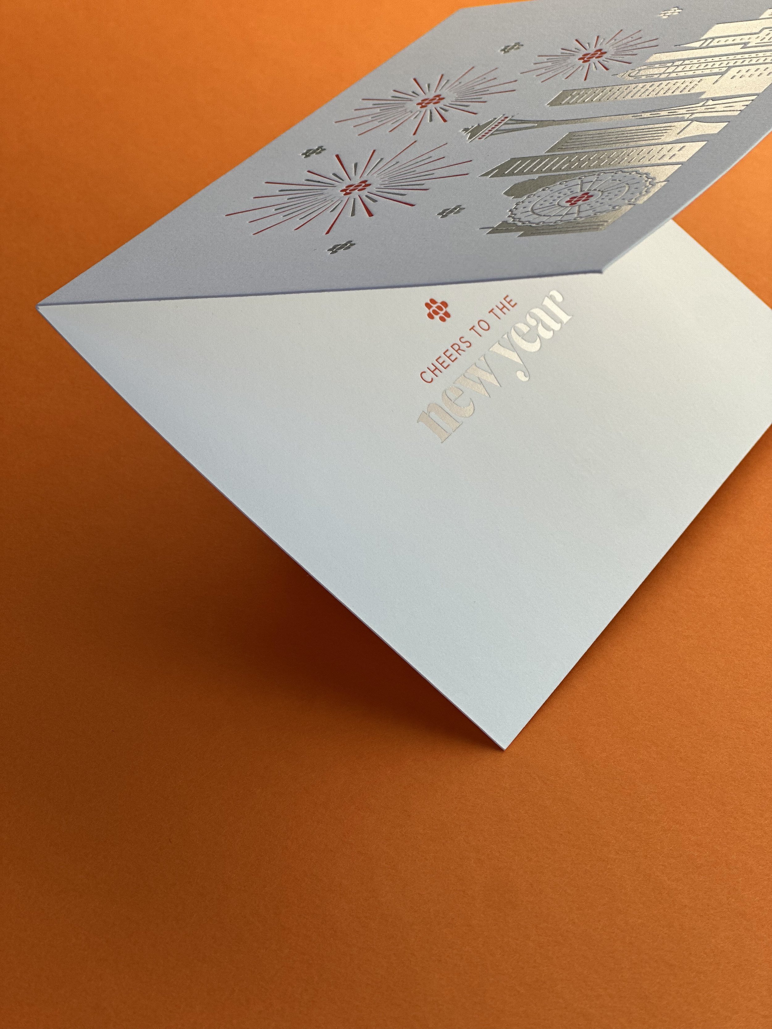 A New Year greeting card with festive fireworks and fireworks designs, with the text "Cheers to the New Year" on a white background, placed on an orange surface.