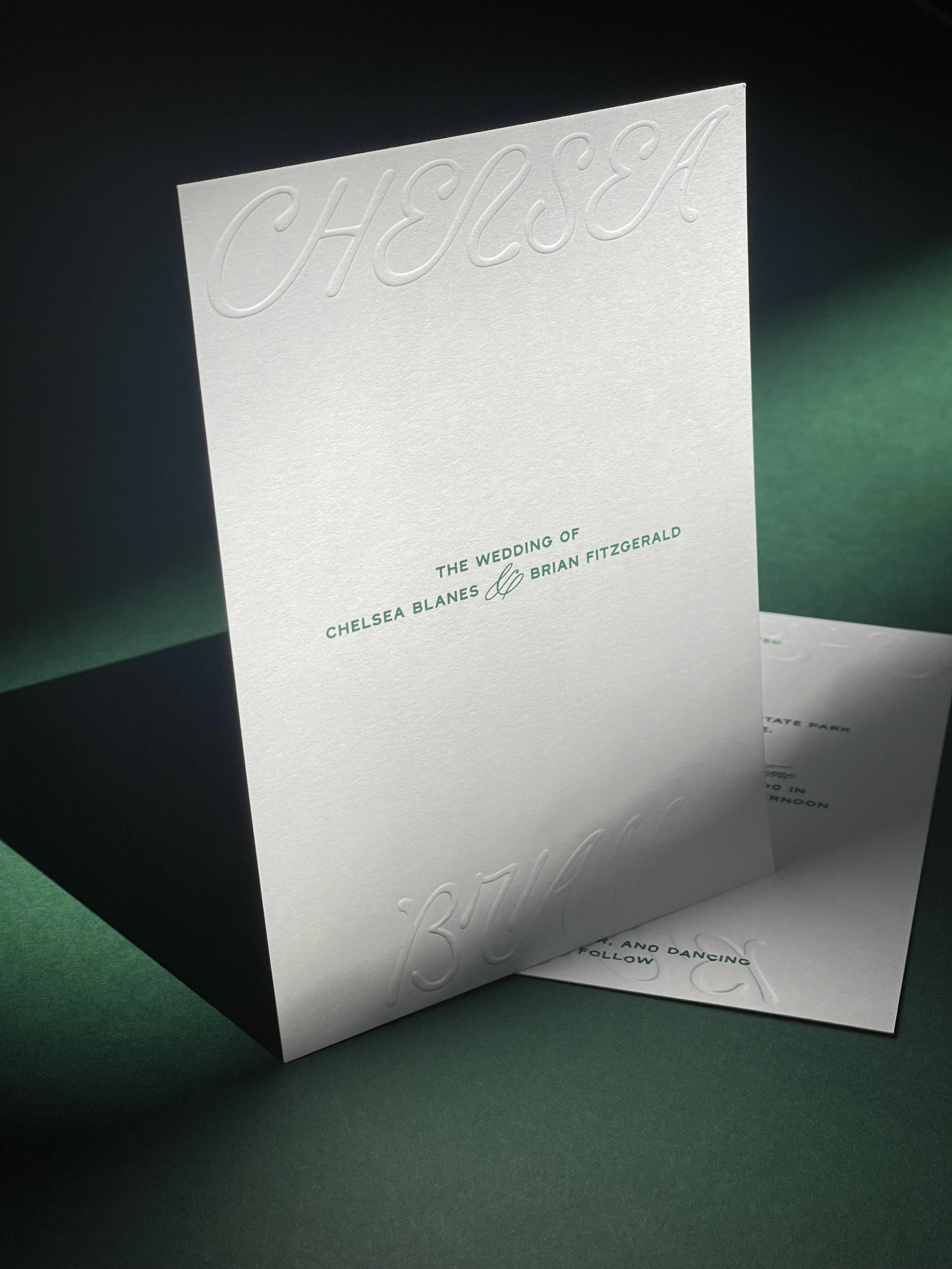 White wedding invitation card with embossed 'CHELSEA' at the top and green text in the center that reads 'The wedding of Chelsea Blanes and Brian Fitzgerald' on a dark green background.