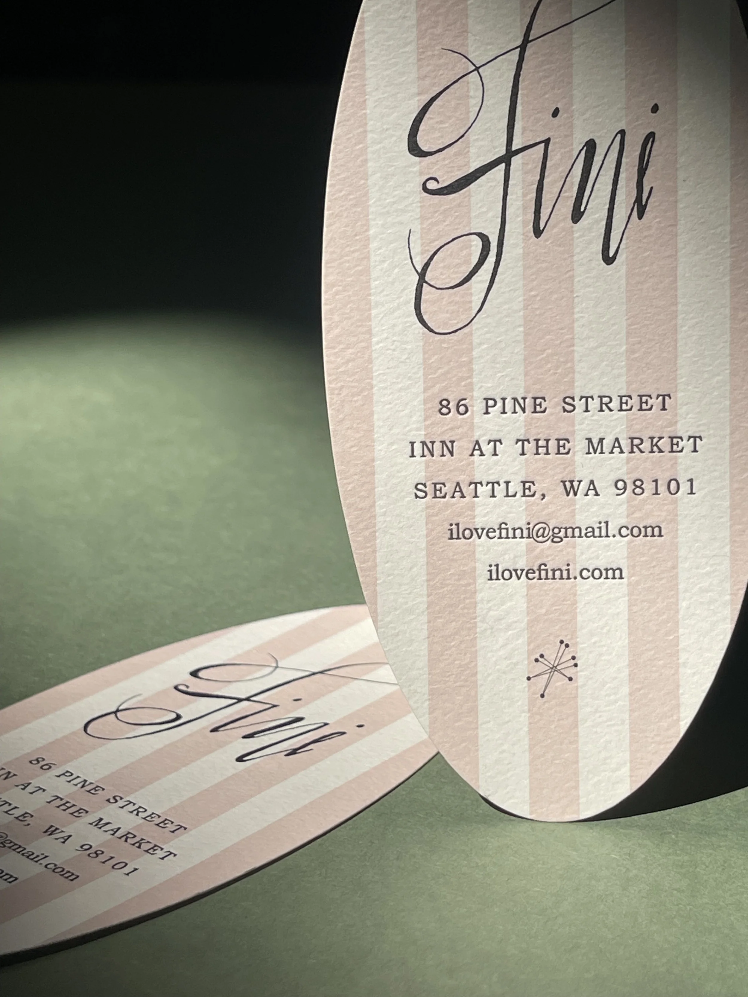 Close-up of a vertically oriented, oval-shaped business card or tag with pink and white vertical stripes. Text includes the name 'Fiini' in cursive, and address details for a location in Seattle, Washington, along with an email address and website.