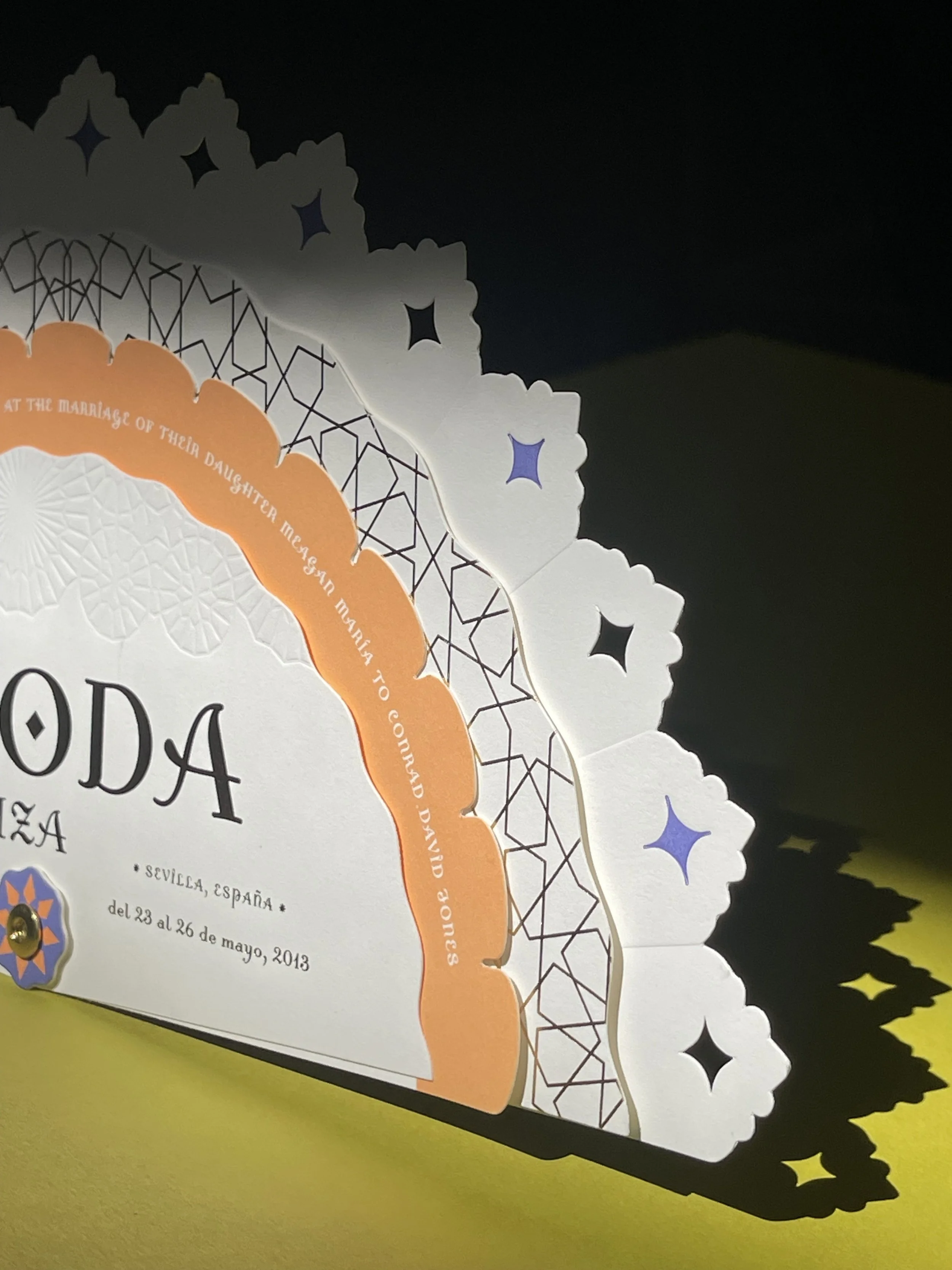 Decorative paper invitation with scalloped edges, floral patterns, and geometric designs in white, black, orange, and purple colors, casting intricate shadows on a yellow surface.