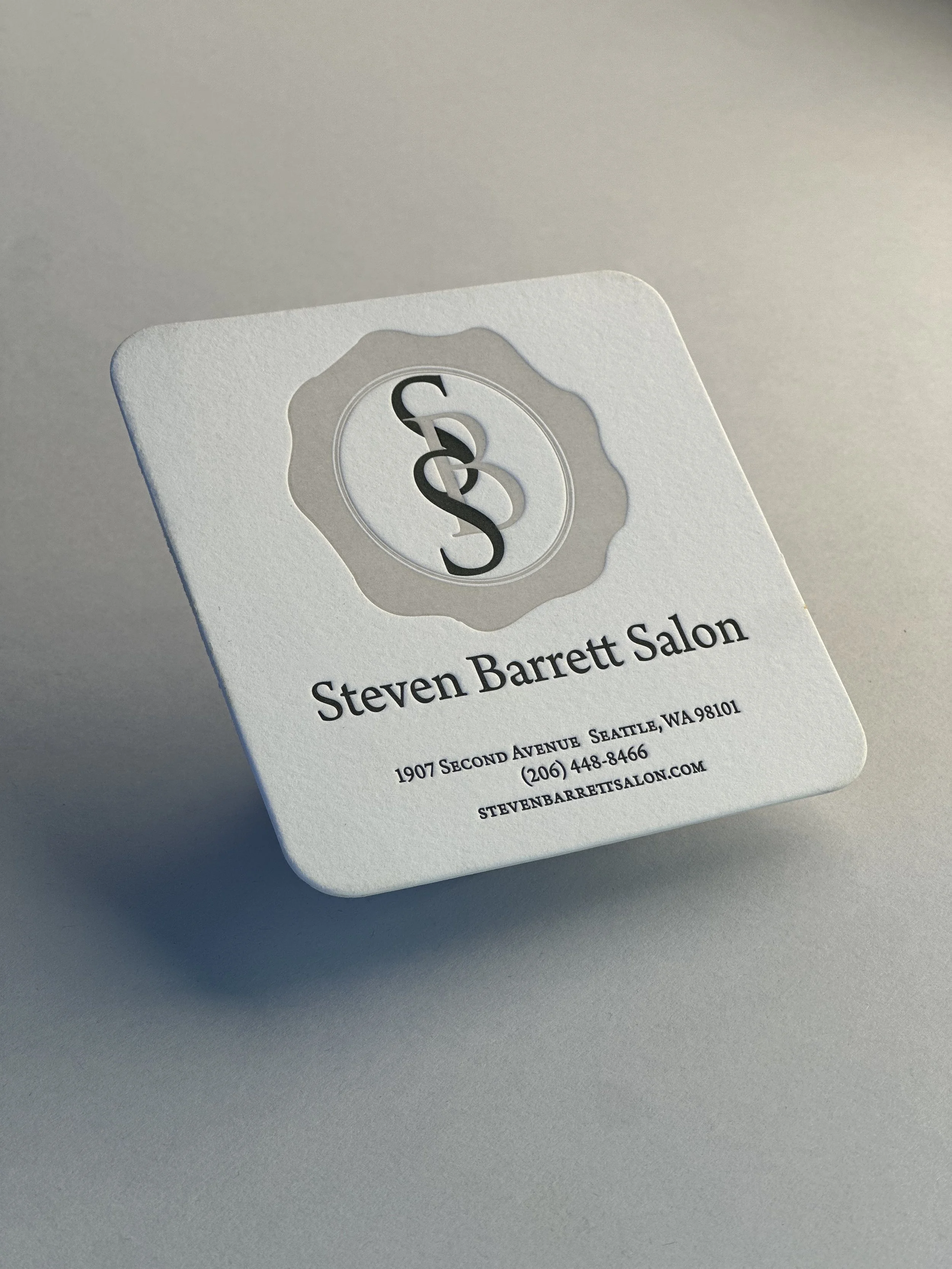 A square business card for Steven Barrett Salon with a logo at the top, the name Steven Barrett Salon in bold, the address 1907 Second Avenue Seattle, WA 98101, phone number (206) 448-8466, and website stevenbarrettsalon.com.