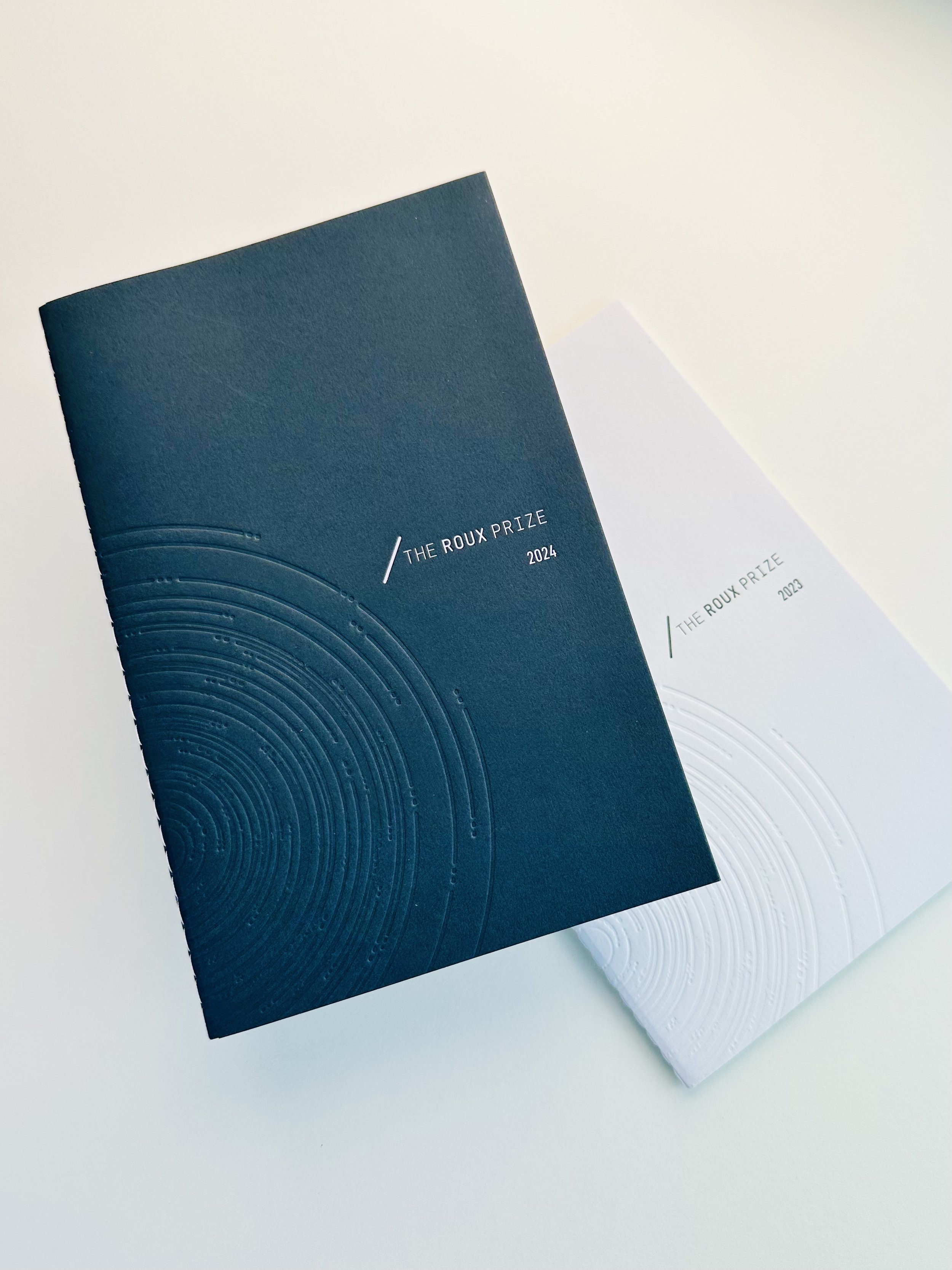Two booklets, one black and one white, labeled 'The Roux Prize' for the years 2024 and 2023 respectively, with embossed circular designs on the covers, placed on a white surface.