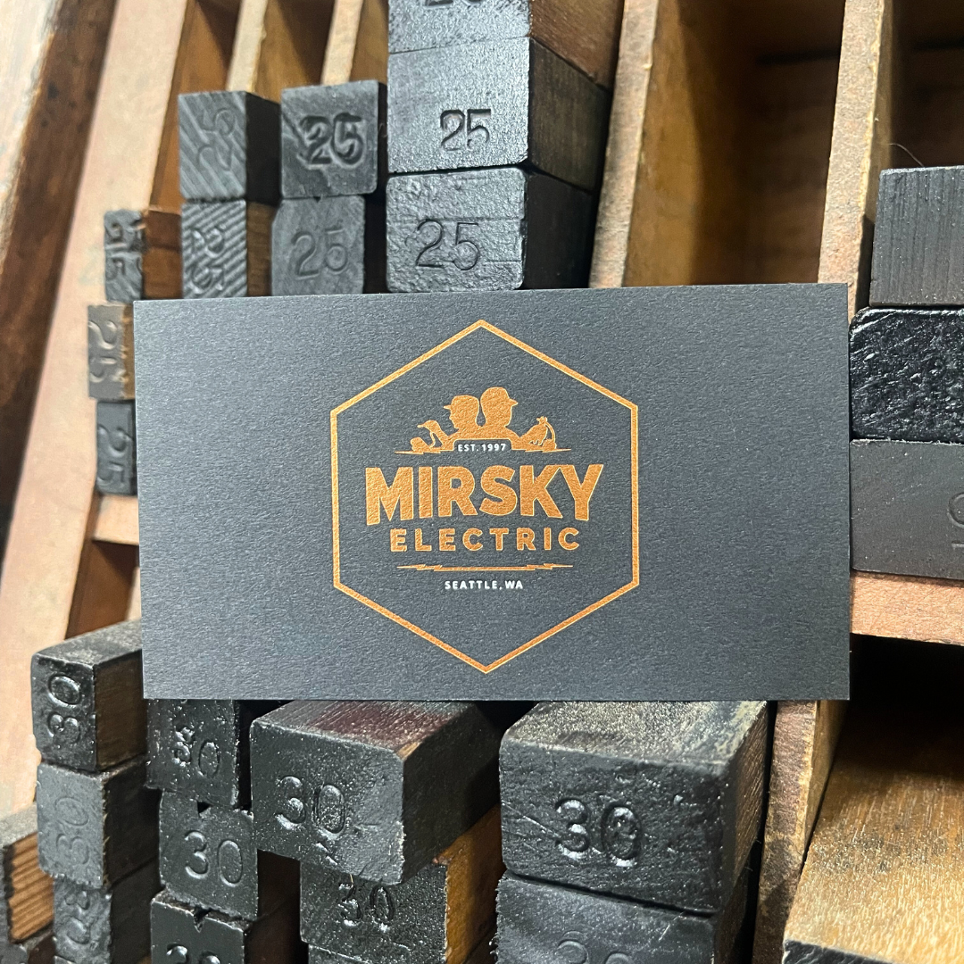 Business card for Mirsky Electric in Seattle, WA, placed on a wooden surface among metal stamps marked with numbers, including 25 and 30.