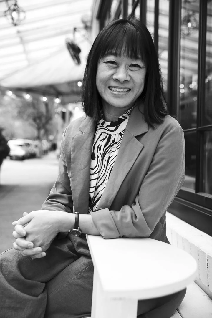 A smiling woman with shoulder-length dark hair sitting outside near a window on a white chair. She is wearing a blazer over a patterned blouse and has a watch on her left wrist. The background features a canopy and parked cars.