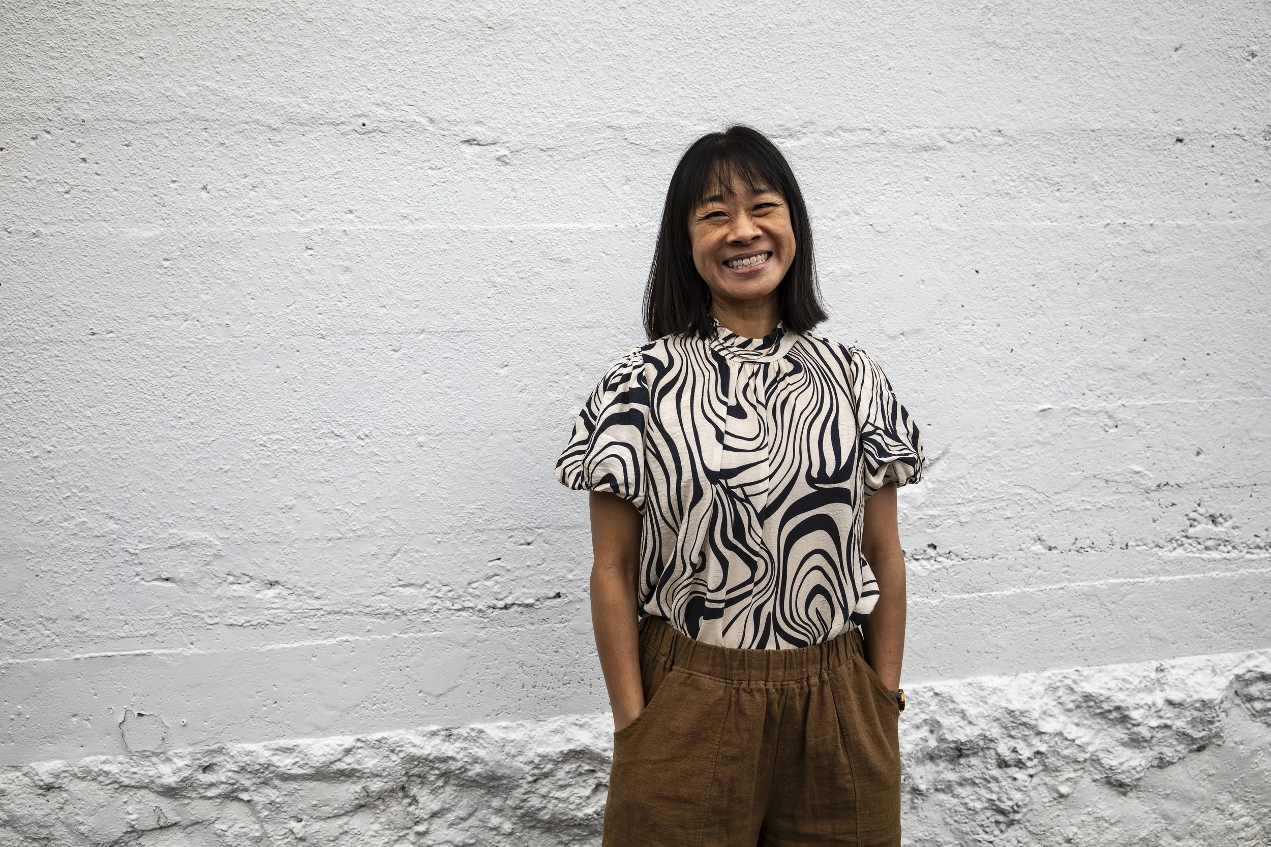 Woman with shoulder-length black hair, smiling, wearing a patterned short-sleeve top and brown pants, standing against a textured white wall.