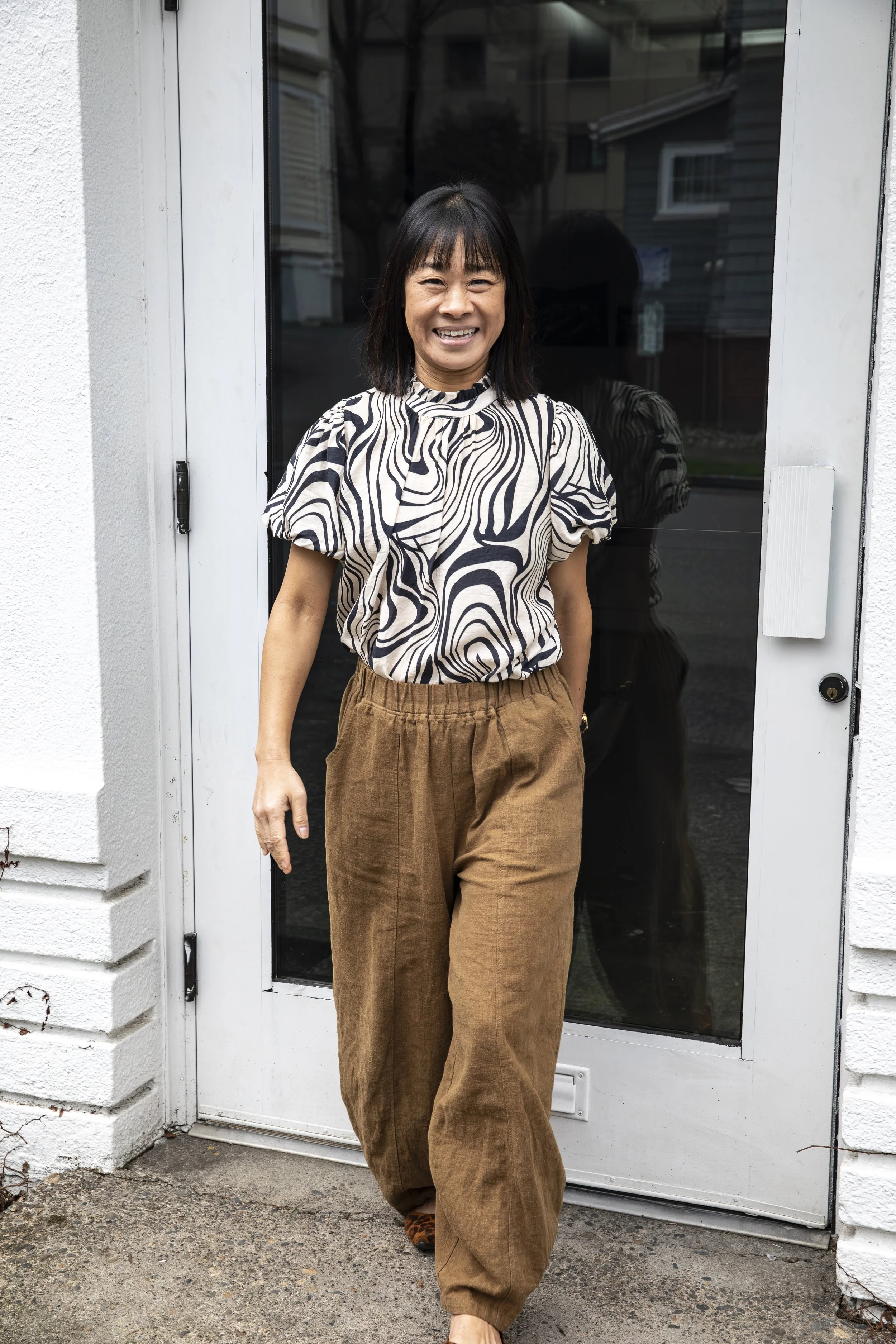 A woman with shoulder-length dark hair, smiling, standing in front of a glass door with a black frame. She is wearing a black and beige patterned blouse and loose brown trousers, with her left hand by her side and her right hand slightly behind her.